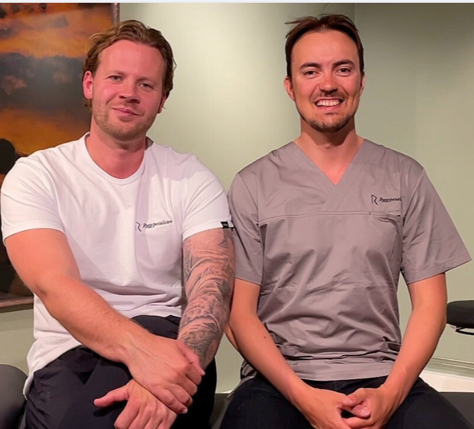 To menn sitter sammen, den ene i hvit t-skjorte og den andre i grå arbeidsuniform, inne i et rom med en grønn vegg. Begge smiler.