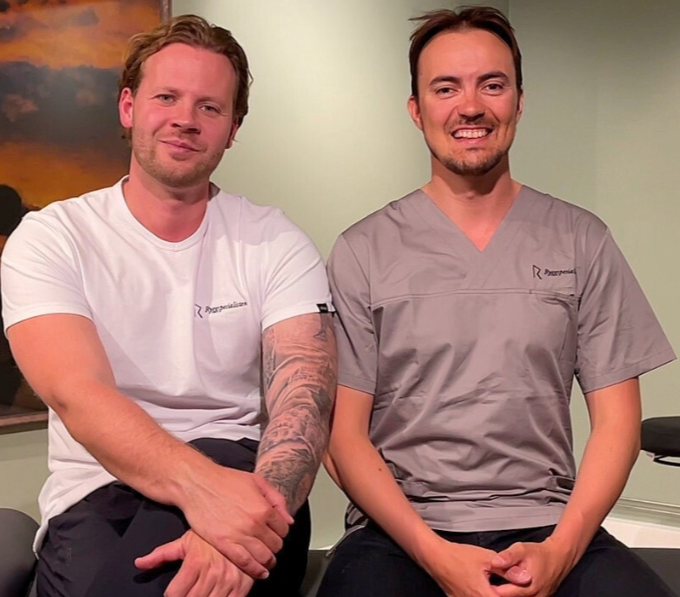 To menn i casual klær sitter sammen og smiler.