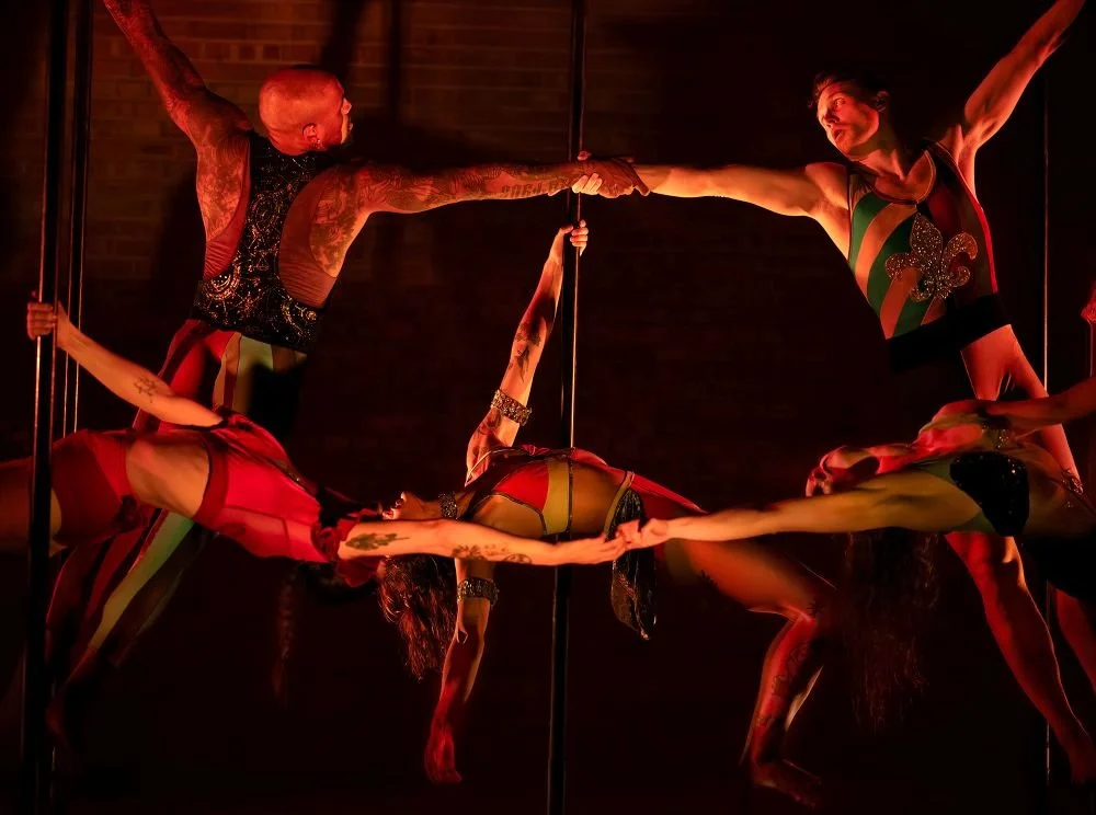 Four female pole dancers performing a coordinated acro-dance routine on a vertical pole in a dimly lit setting.
