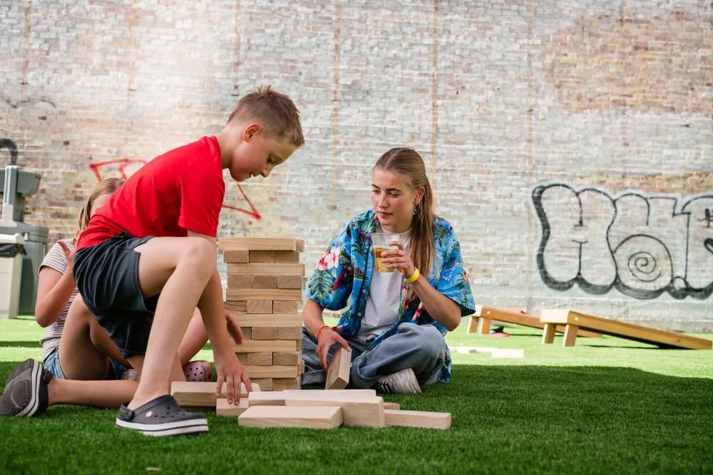 Children playing a giant Jenga game outdoors on artificial grass with a brick wall background and graffiti.