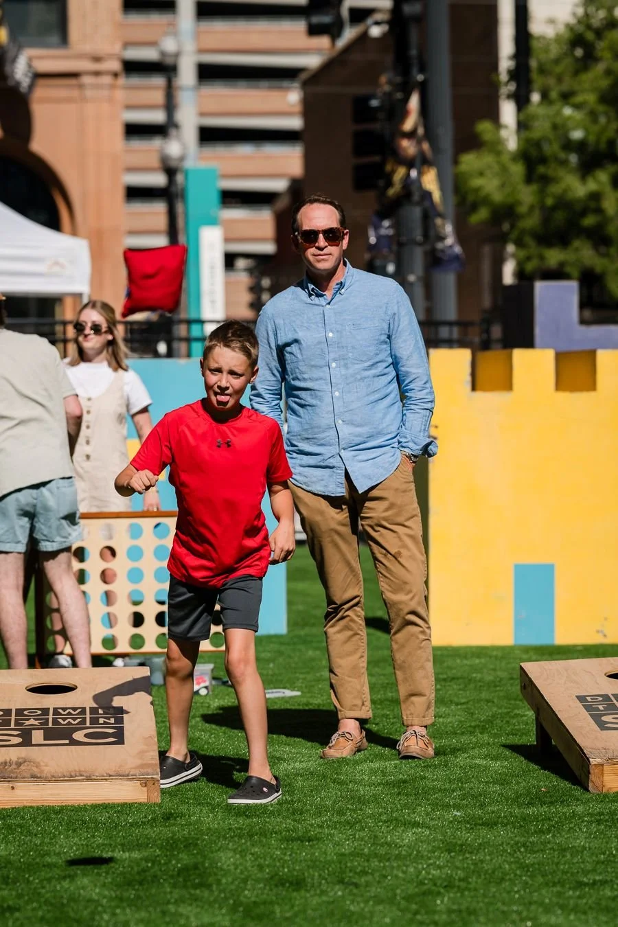 A young boy in a red shirt and black shorts playing a ring toss game outdoors on green grass, with a man in sunglasses and a blue shirt standing nearby, and other people in the background during a sunny day.