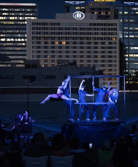 A nighttime outdoor performance with a group of acrobats on a stage with a metal frame, city buildings in the background, and audience watching in the foreground.