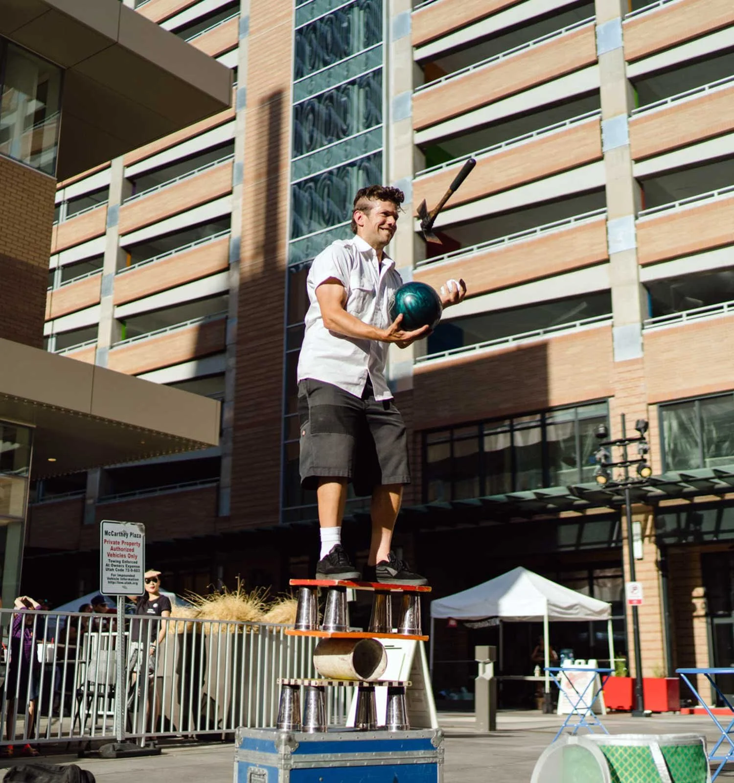 Man standing on a structure of items while juggling a bowling ball and hatchet.