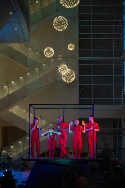 Five performers in red outfits stand on a platform in an indoor venue with modern architecture, large balconies, and spherical hanging lights.