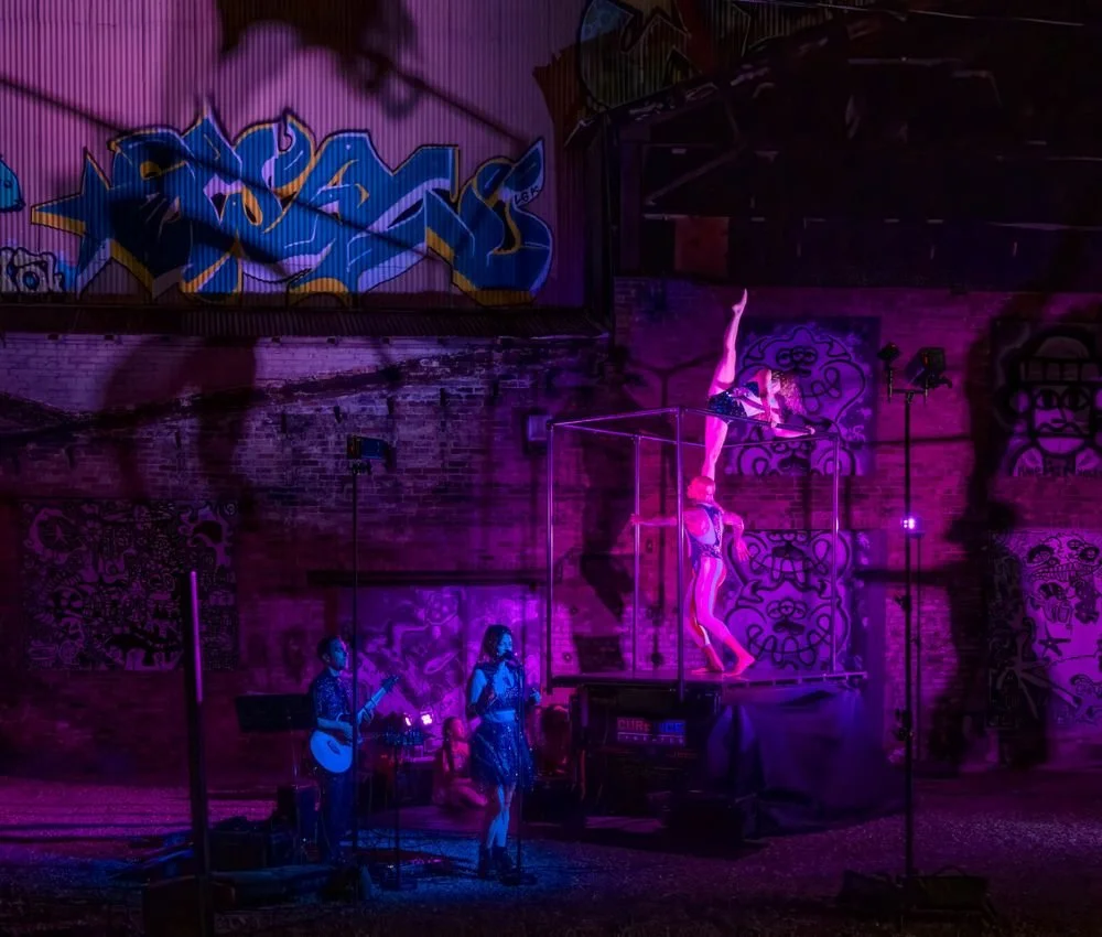 A performance scene in a dimly lit room with purple and pink lights, featuring two singers and two acrobats. One acrobat is performing a handstand on top of a platform, while another is hanging upside down, supported by a performer.