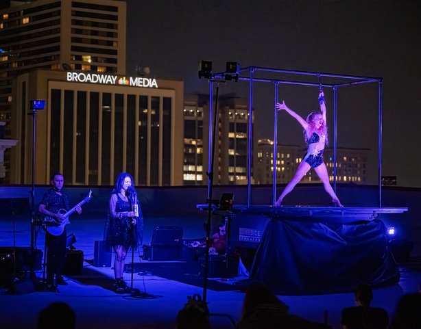 A nighttime outdoor performance on a rooftop stage with city buildings in the background. There is a dancer in a sparkly outfit on a elevated platform, striking a pose with arms outstretched. Two musicians, a guitarist and a singer, are performing be