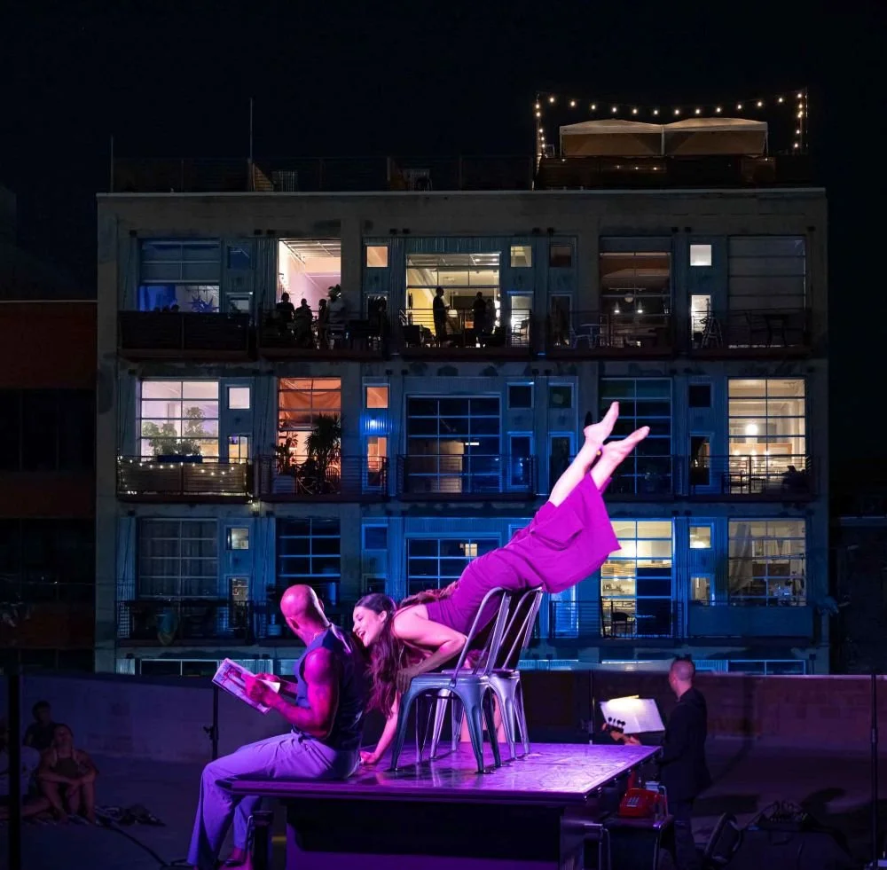 A woman in purple attire performs a balancing act on a chairs stage with a man reading a book seated beside her. An audience watches as a performer on the right reads sheet music. The background features a building with lit windows and an outdoor seating area with string lights.