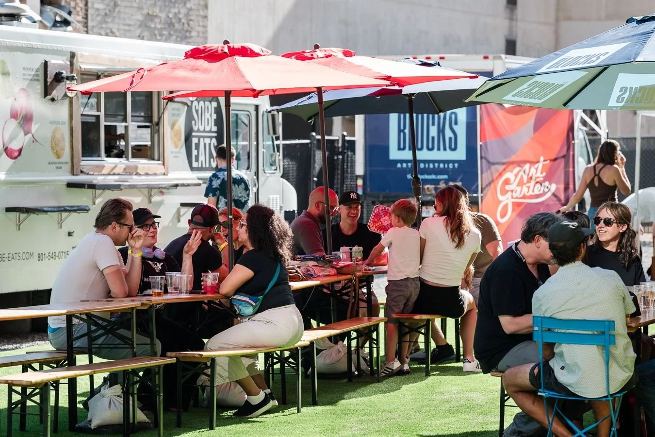People sitting and socializing at picnic tables with umbrellas at an outdoor food truck park.