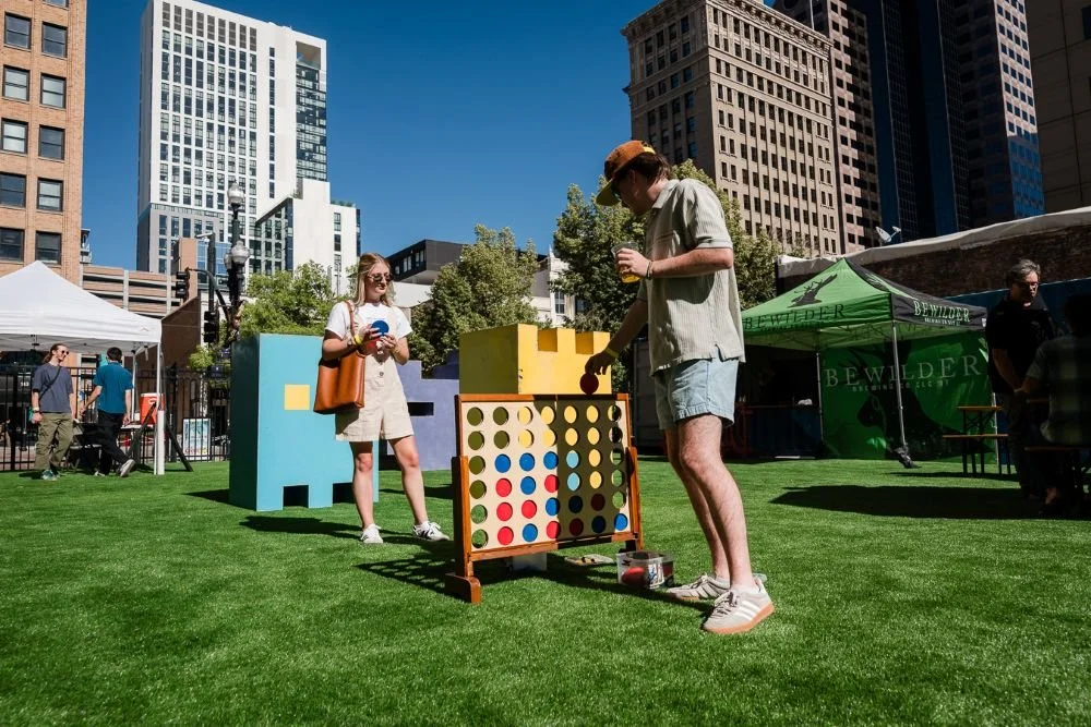 People playing giant Connect Four game at an outdoor festival in an urban park with tall buildings in the background.