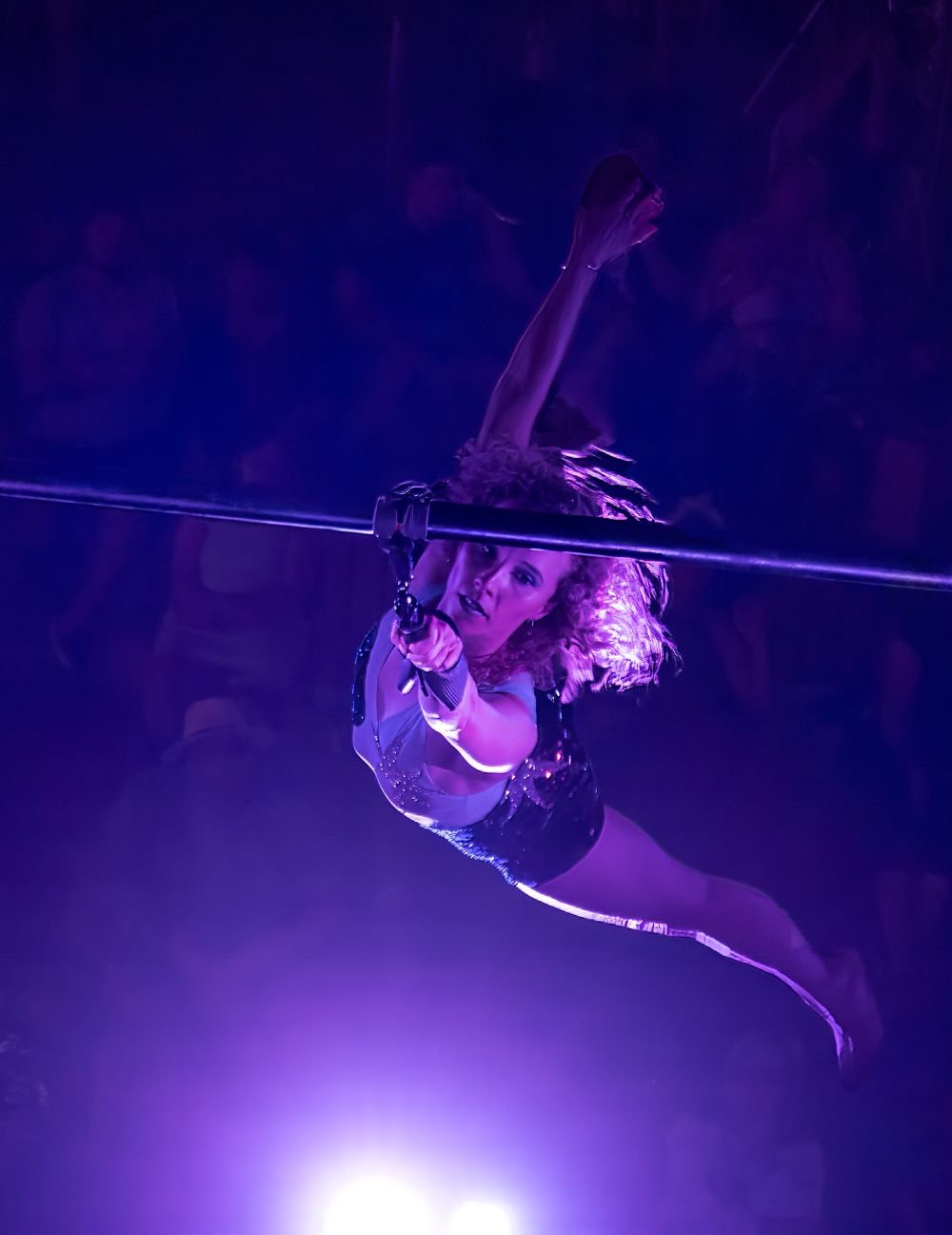 A female performer with curly hair hanging from a trapeze, illuminated by purple stage lights, performing an aerial act.