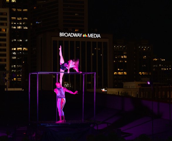 Two performers practicing acrobatics on a lit-up trapeze on a rooftop at night, with city buildings and a Broadway Media sign in the background.