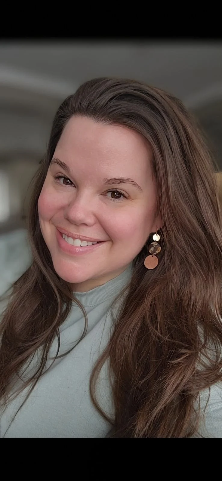 Smiling woman with long brown hair wearing earrings and a gray top.