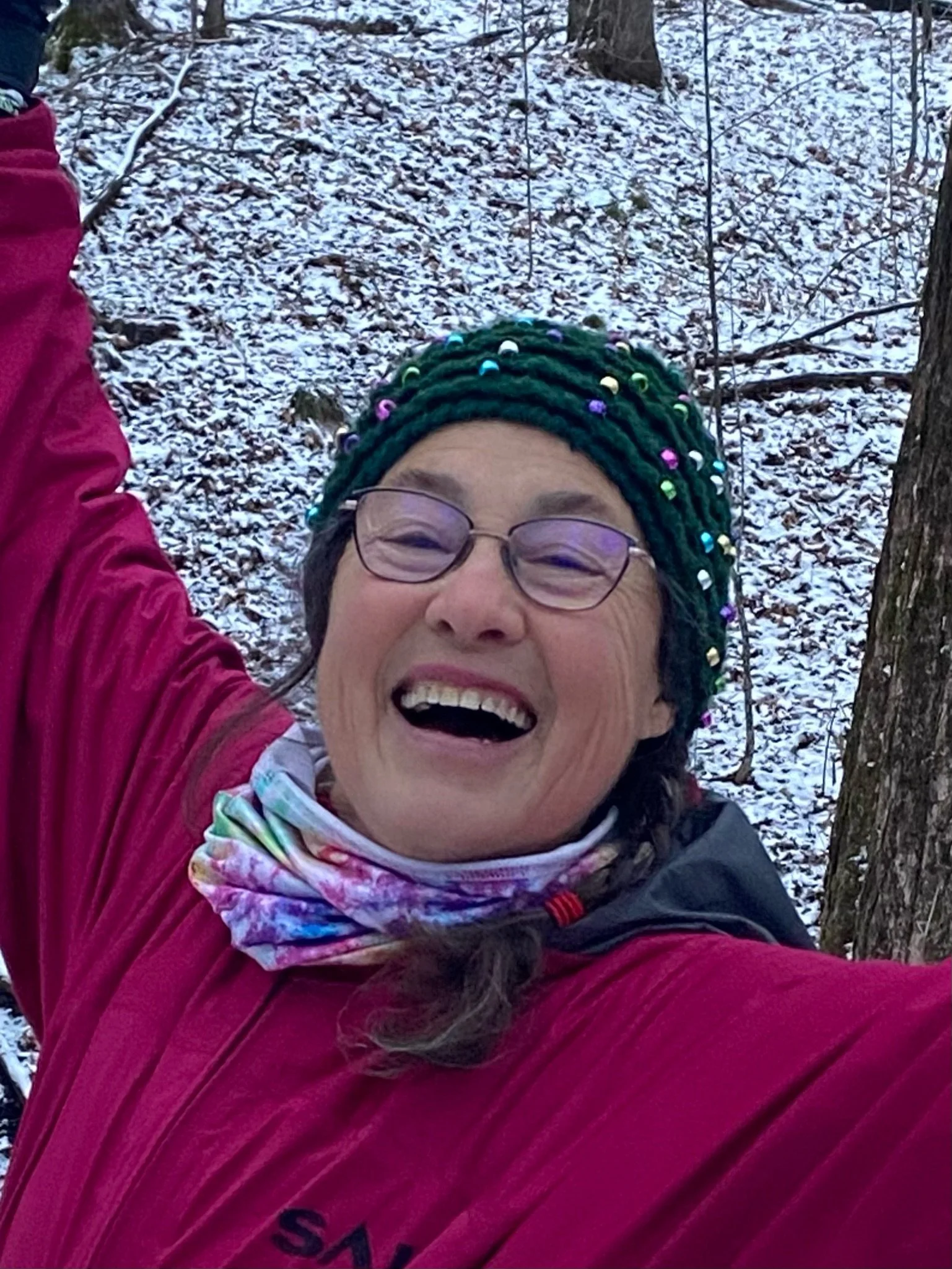 A woman outdoors in winter, wearing a colorful scarf, a beaded knit hat, and a red jacket, smiling with arms raised against a snowy background.