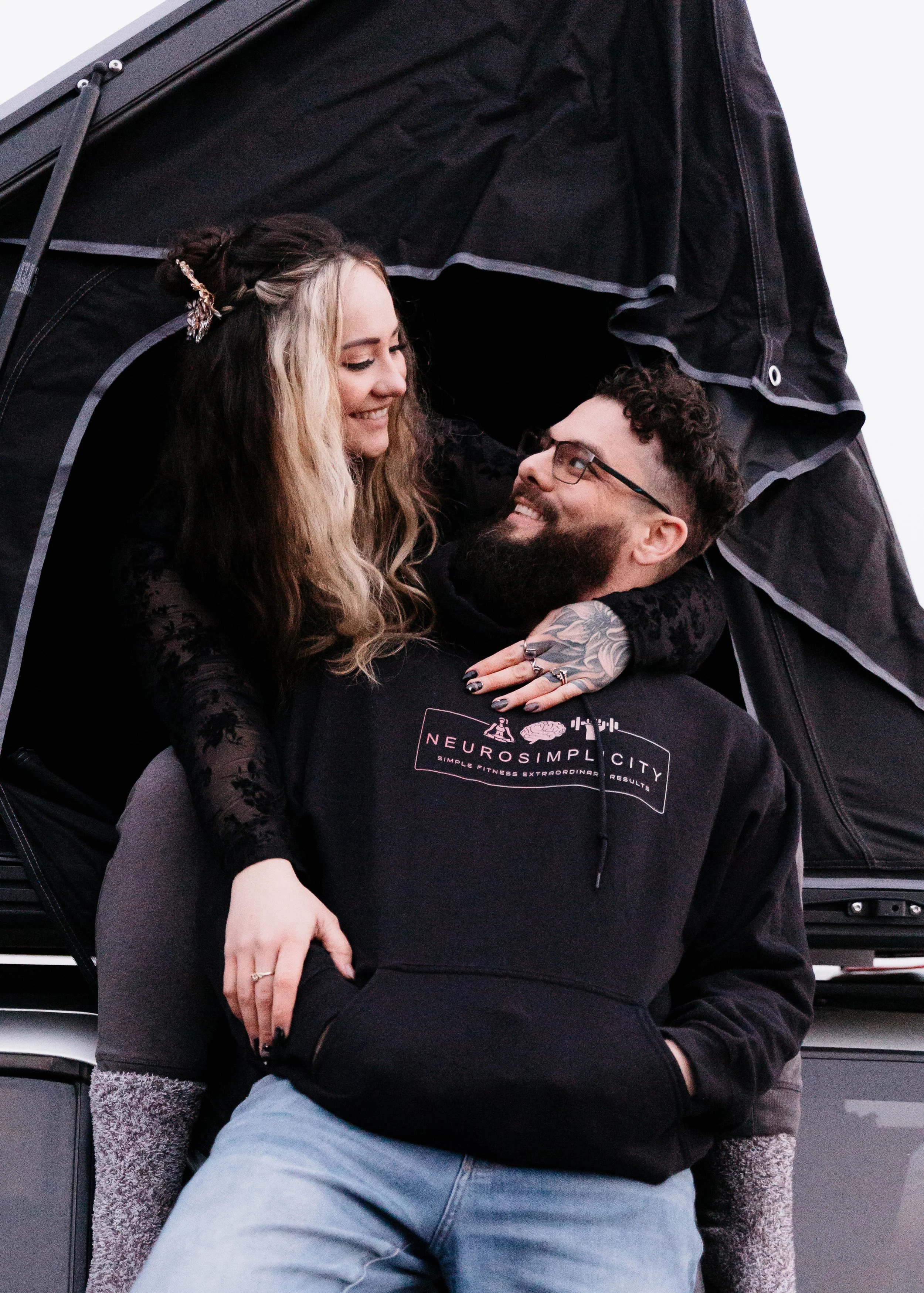 A smiling couple sitting close together, with the woman sitting on the man's lap under a black tent or canopy. The woman has long, wavy blonde hair, with a hairpin, and is wearing a black, long-sleeved lace top. The man has a beard, glasses, and is wearing a black hoodie with the logo "NeuroSimplicity" on it. They are looking at each other affectionately.