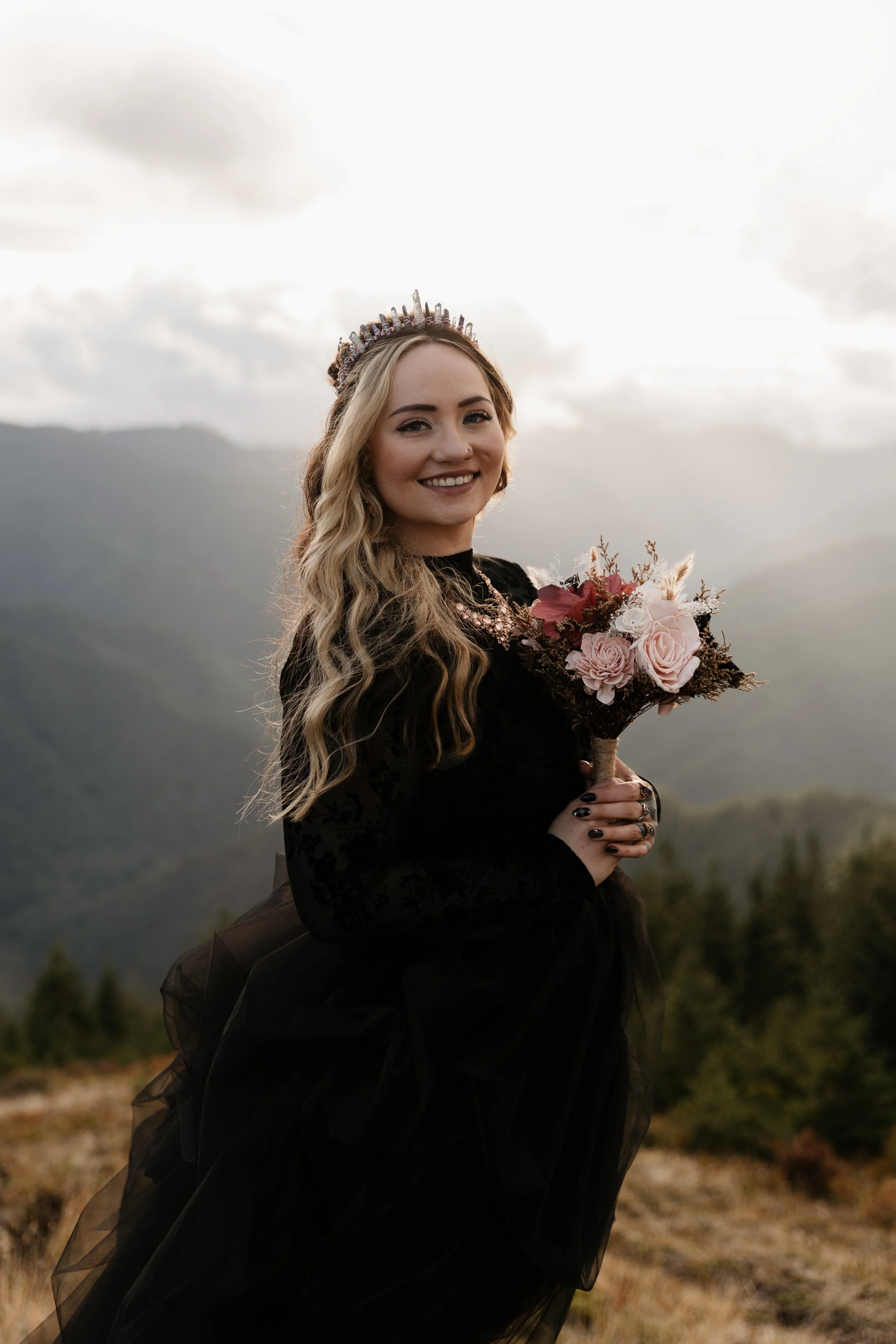 A woman with long blonde hair holding a bouquet of pink and white flowers, wearing a black dress and a crown, standing outdoors with mountains and cloudy sky in the background.