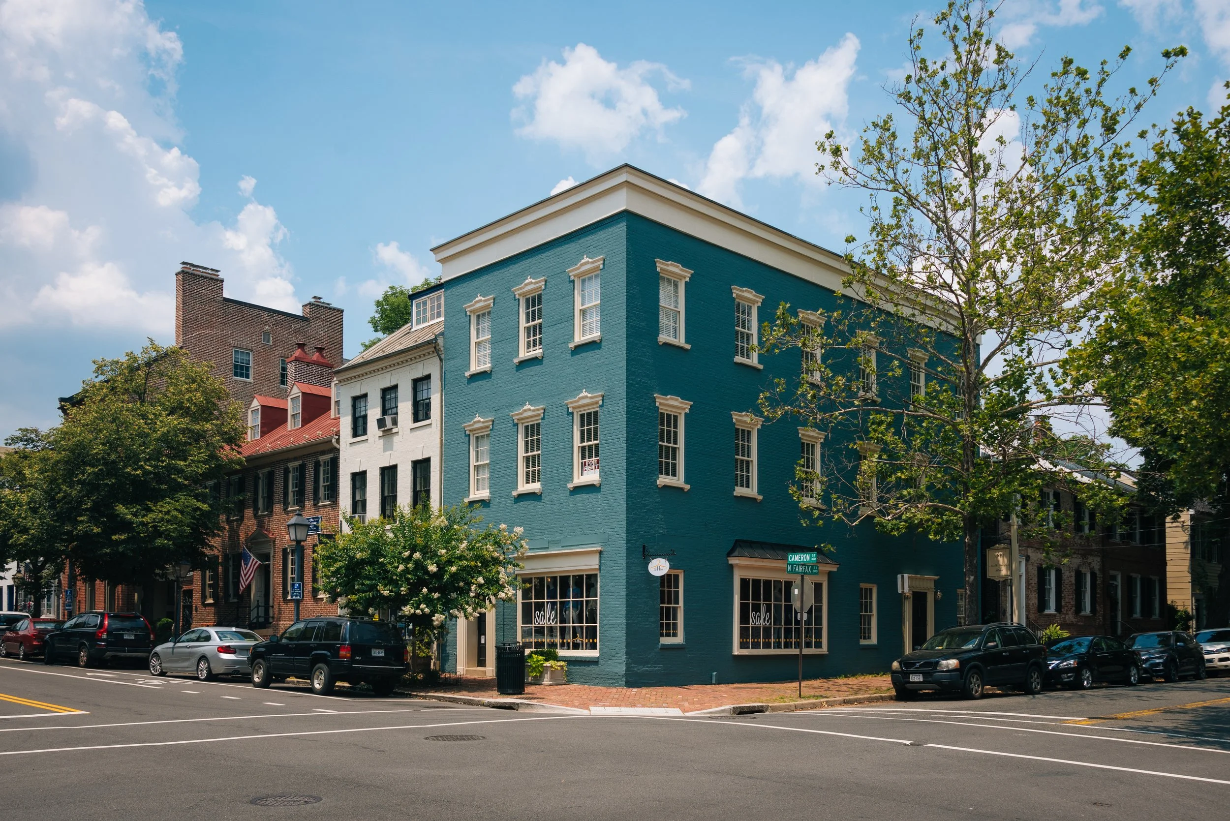 A corner building painted blue with white trim, located on a street with parked cars and trees. Blue sky with clouds.