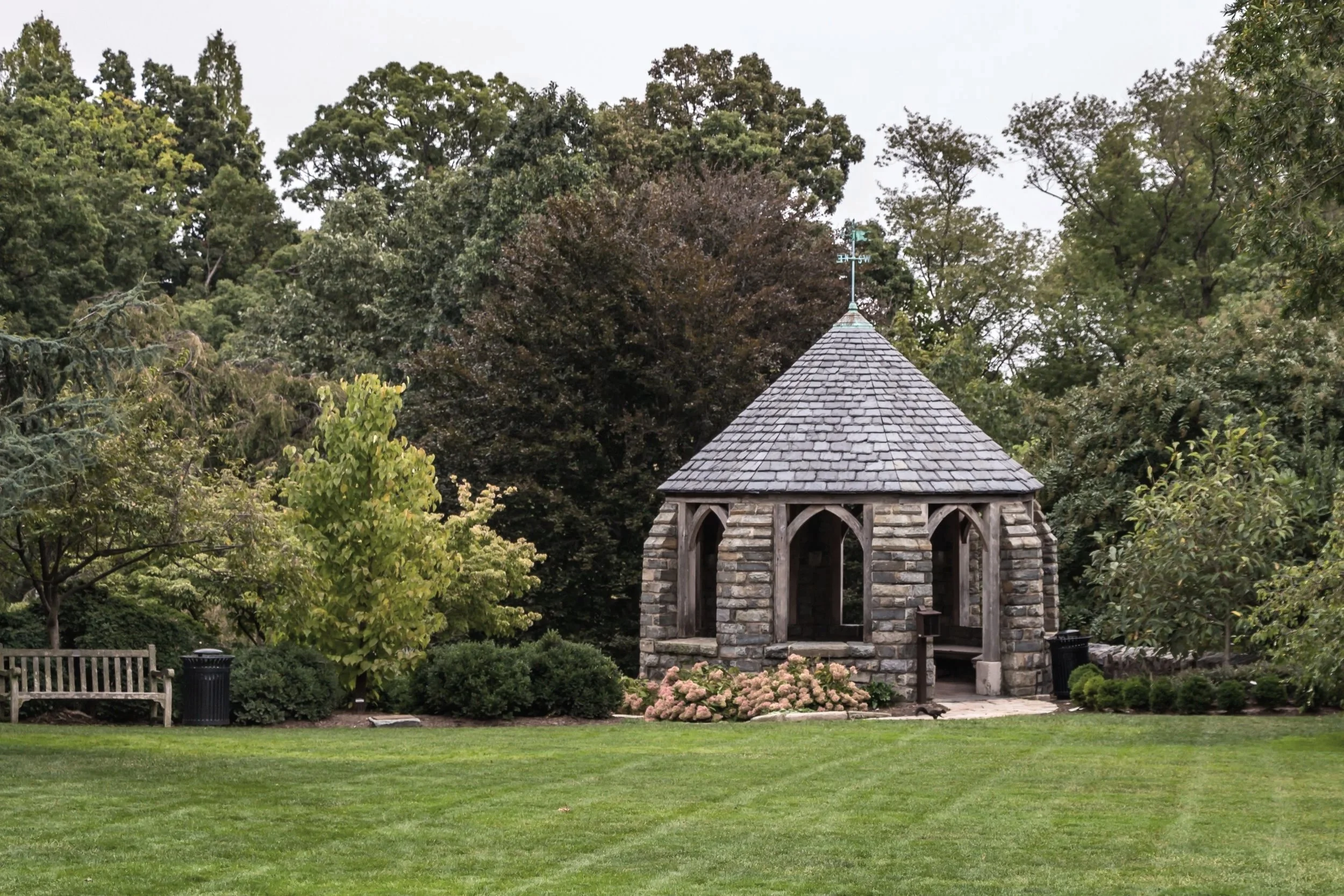A small stone pavilion with a pointed roof, situated in a lush park surrounded by trees and bushes, with a well-maintained lawn in the foreground.
