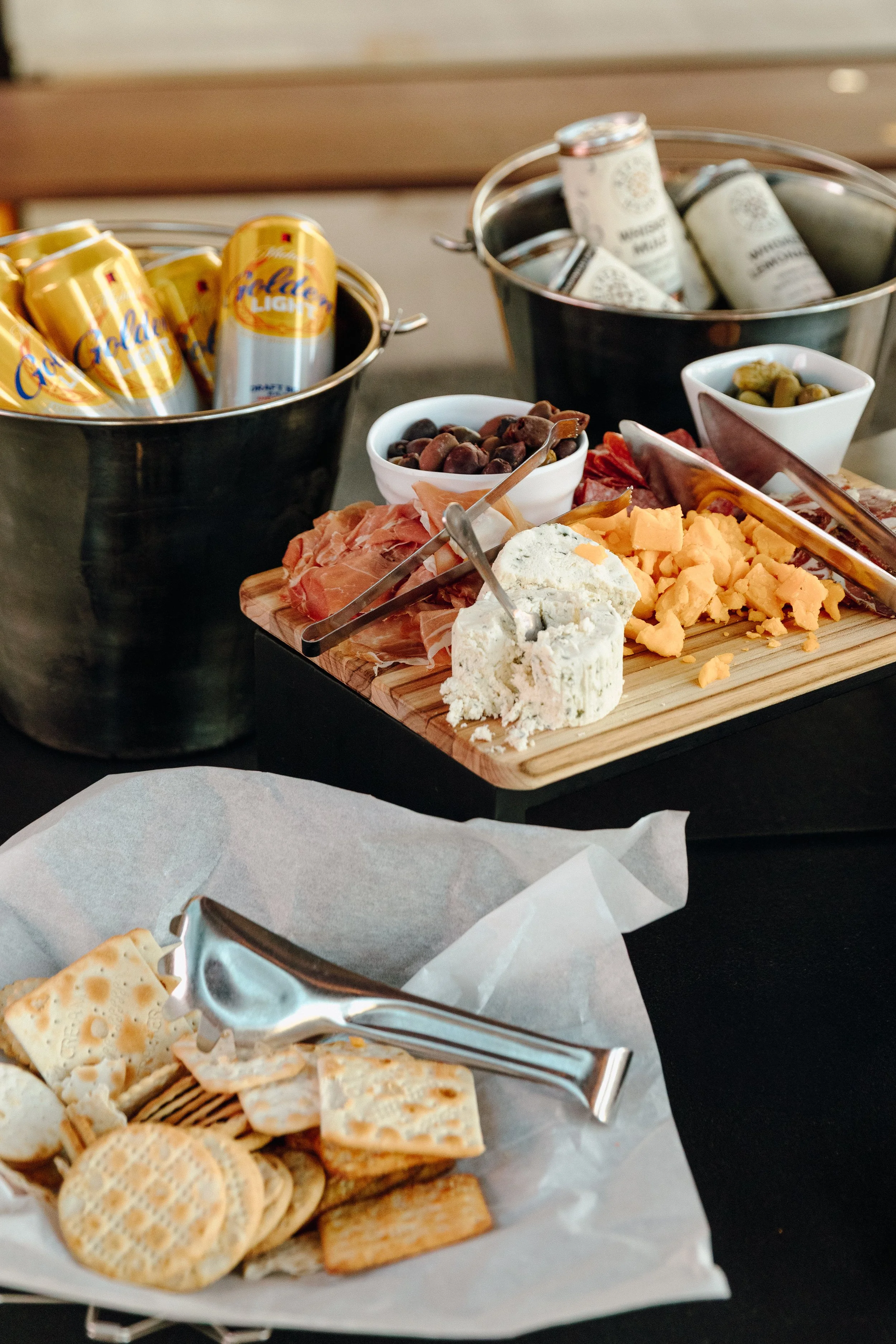 Appetizers at a casual private event at North Loop Green, Minneapolis with charcuterie board with cheese, prosciutto, and olives; two buckets of canned beverages and a basket of crackers in the background.