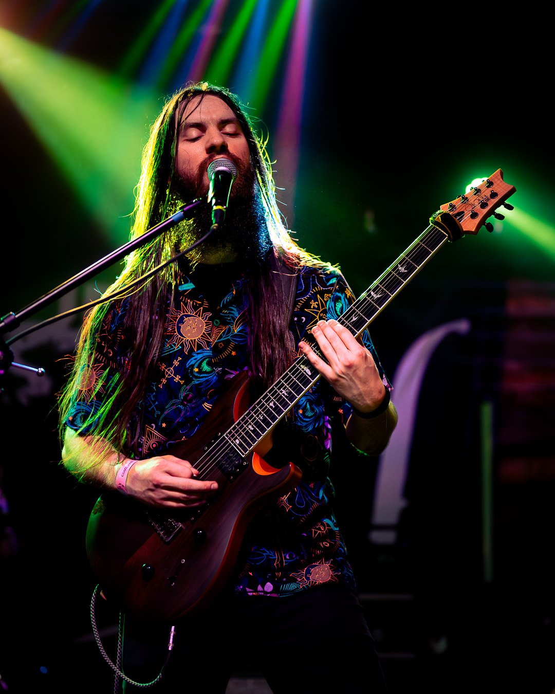 A male musician with long hair and beard singing into a microphone and playing an electric guitar on stage under vibrant green and purple lights.