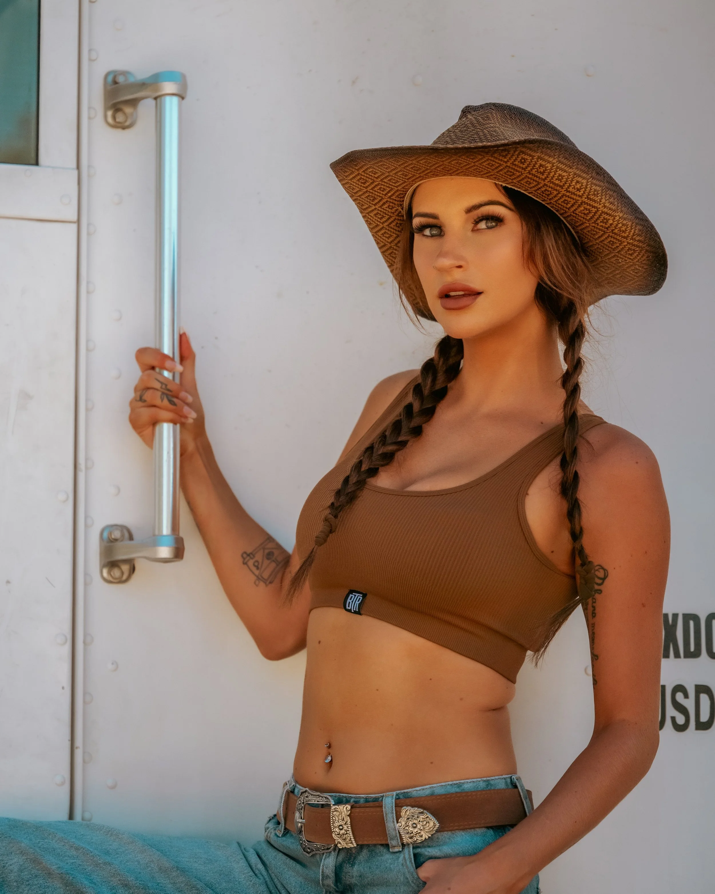Young woman with braided hair wearing a wide-brimmed hat, tank top, jeans, and a belt, standing next to a white wall or truck door.