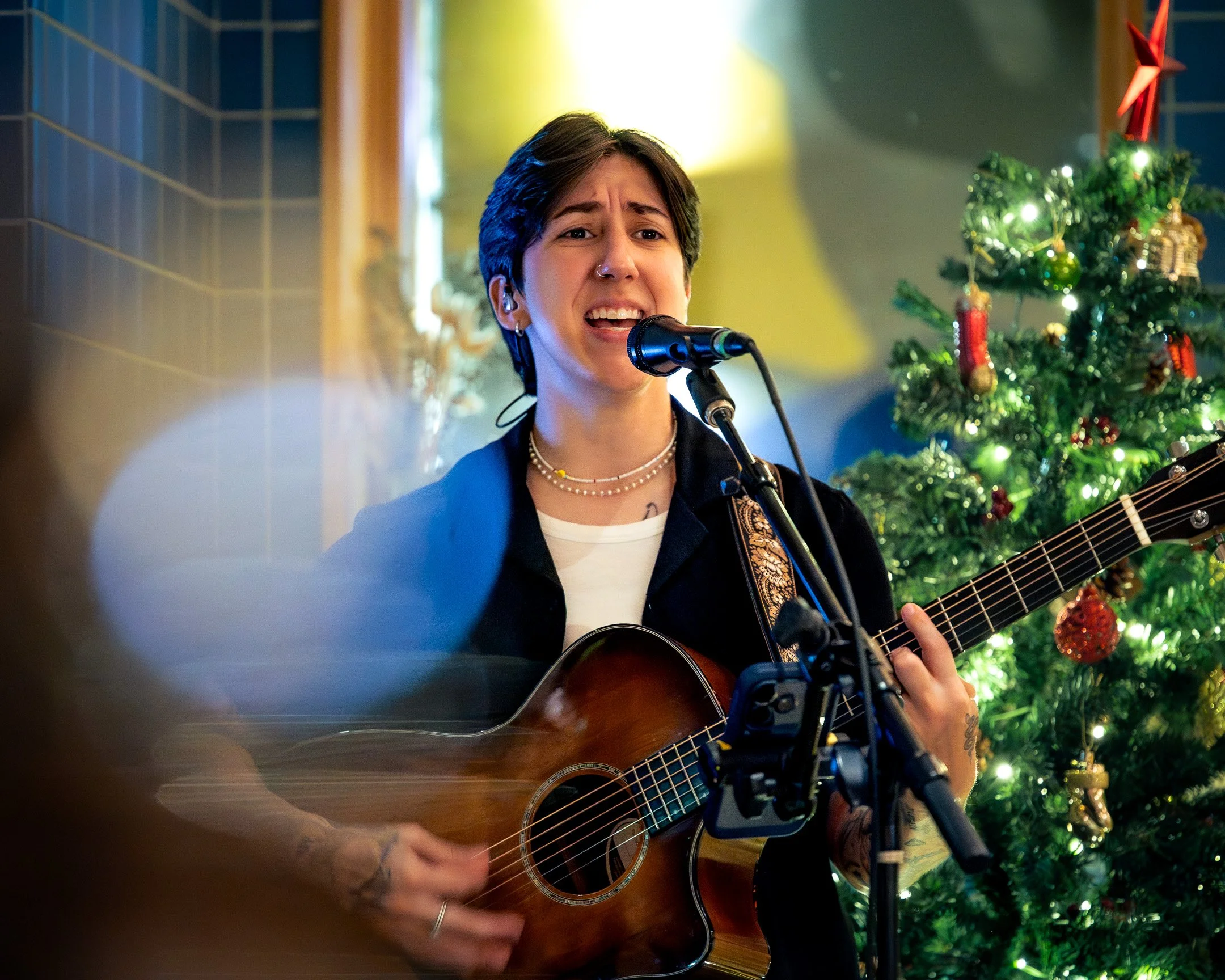 A person with short dark hair, wearing a black jacket and multiple necklaces, is singing into a microphone while playing an acoustic guitar. They are in front of a decorated Christmas tree with ornaments. The background has a yellow lampshade and a blue-tiled wall.