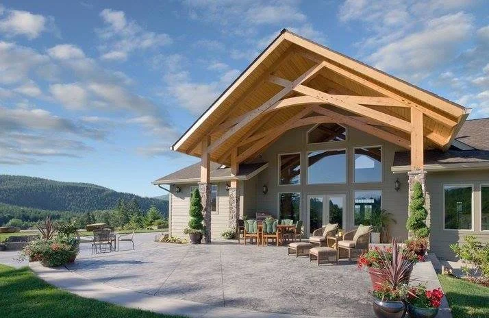 Exterior view of a modern house with a large wooden porch and outdoor seating area, surrounded by a landscaped yard and mountains in the background.