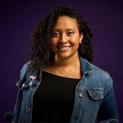 Ash is a black woman with shoulder length black curly hair. She is wearing a black top and a blue denim jacket.