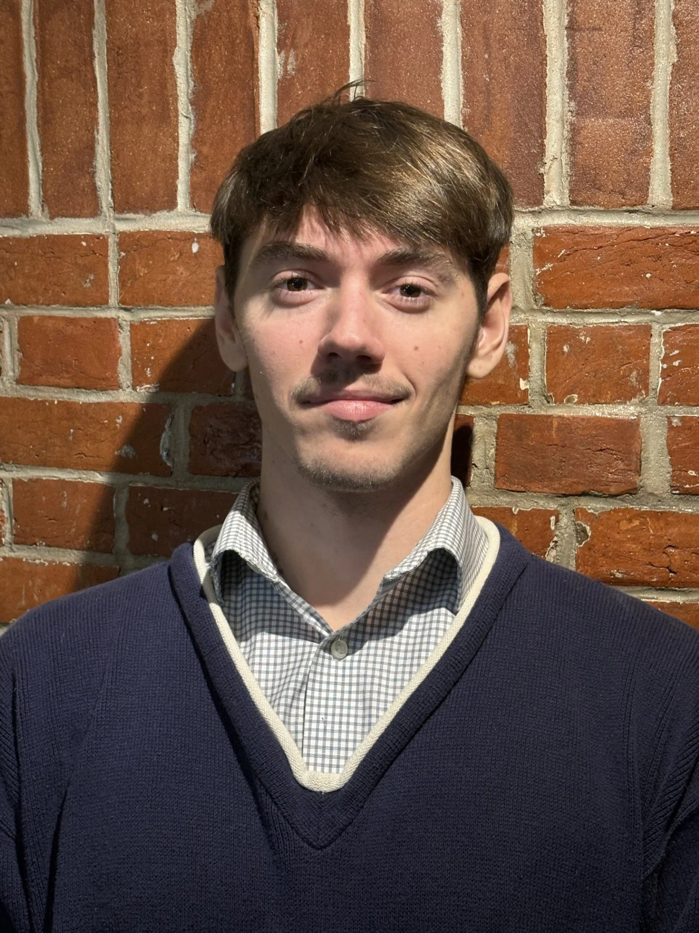 A young woman smiling at the camera at a restaurant or outdoor dining area, wearing a black sleeveless top and a necklace with a flower pendant.