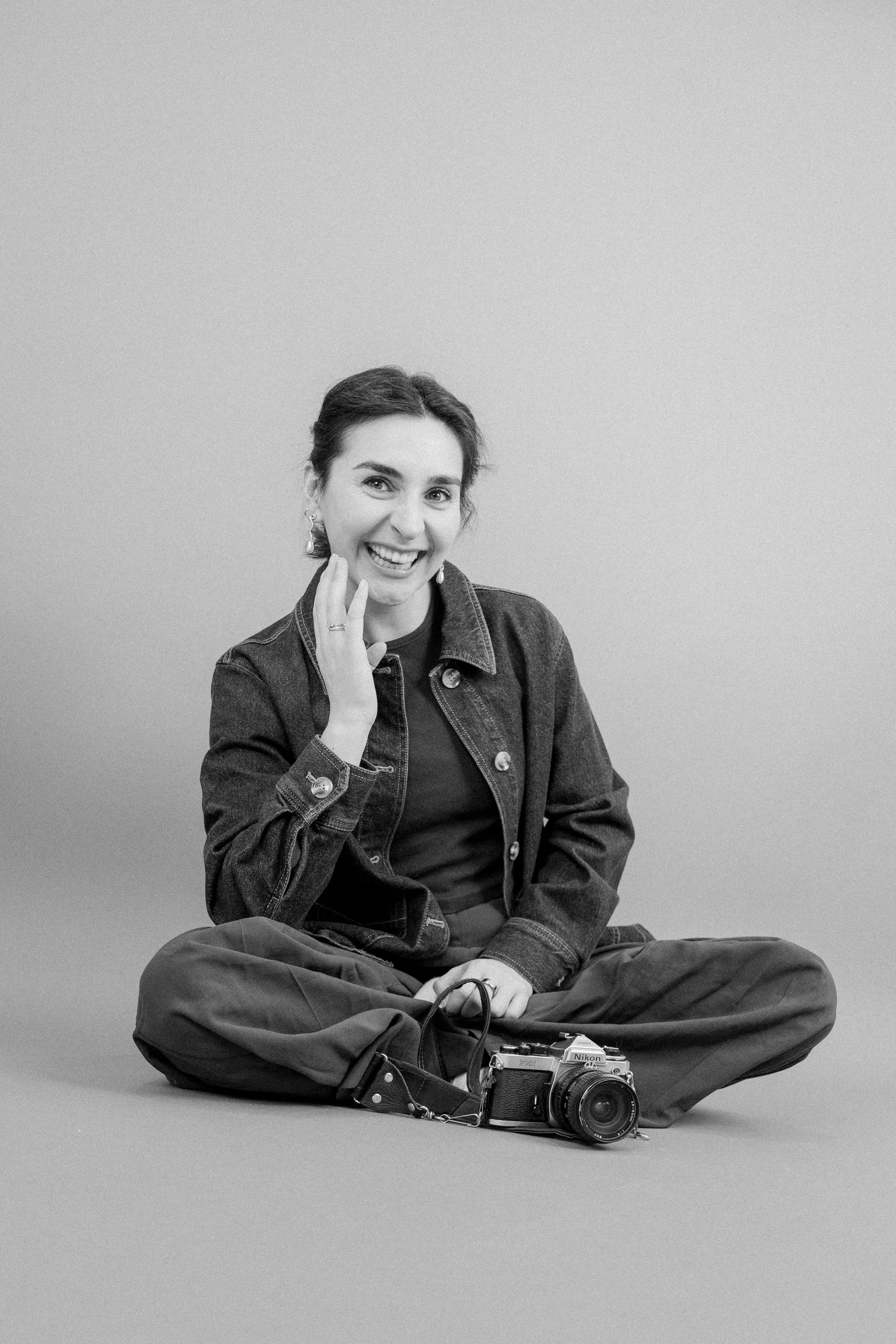 Svart-hvitt bilde av en smilende kvinne som sitter krysset ben og har en Nikon-kamera foran seg.