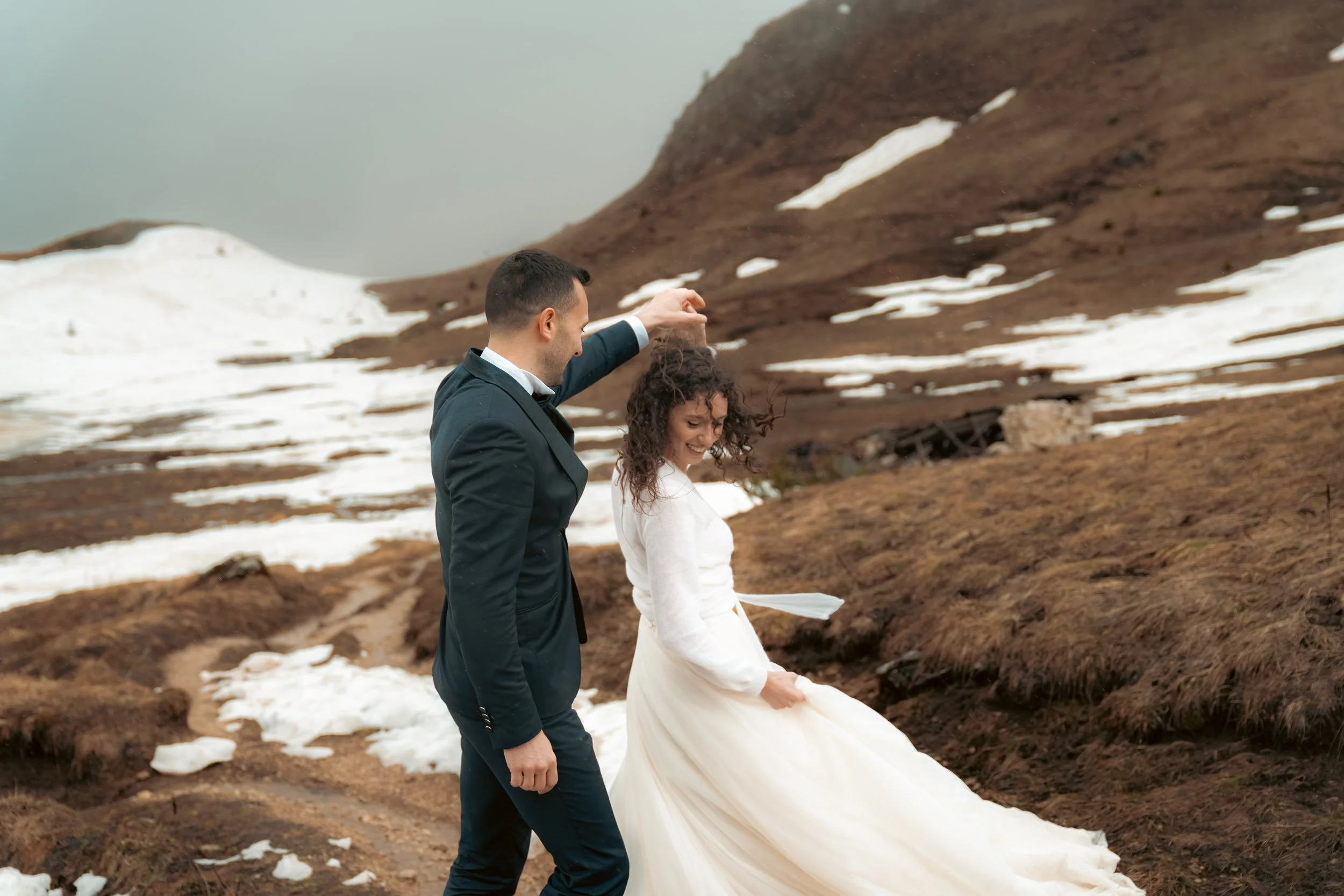 Et livlag, brud og brudgom danser på snødekte fjellstier med brunt landskap og snøfjell i bakgrunnen.