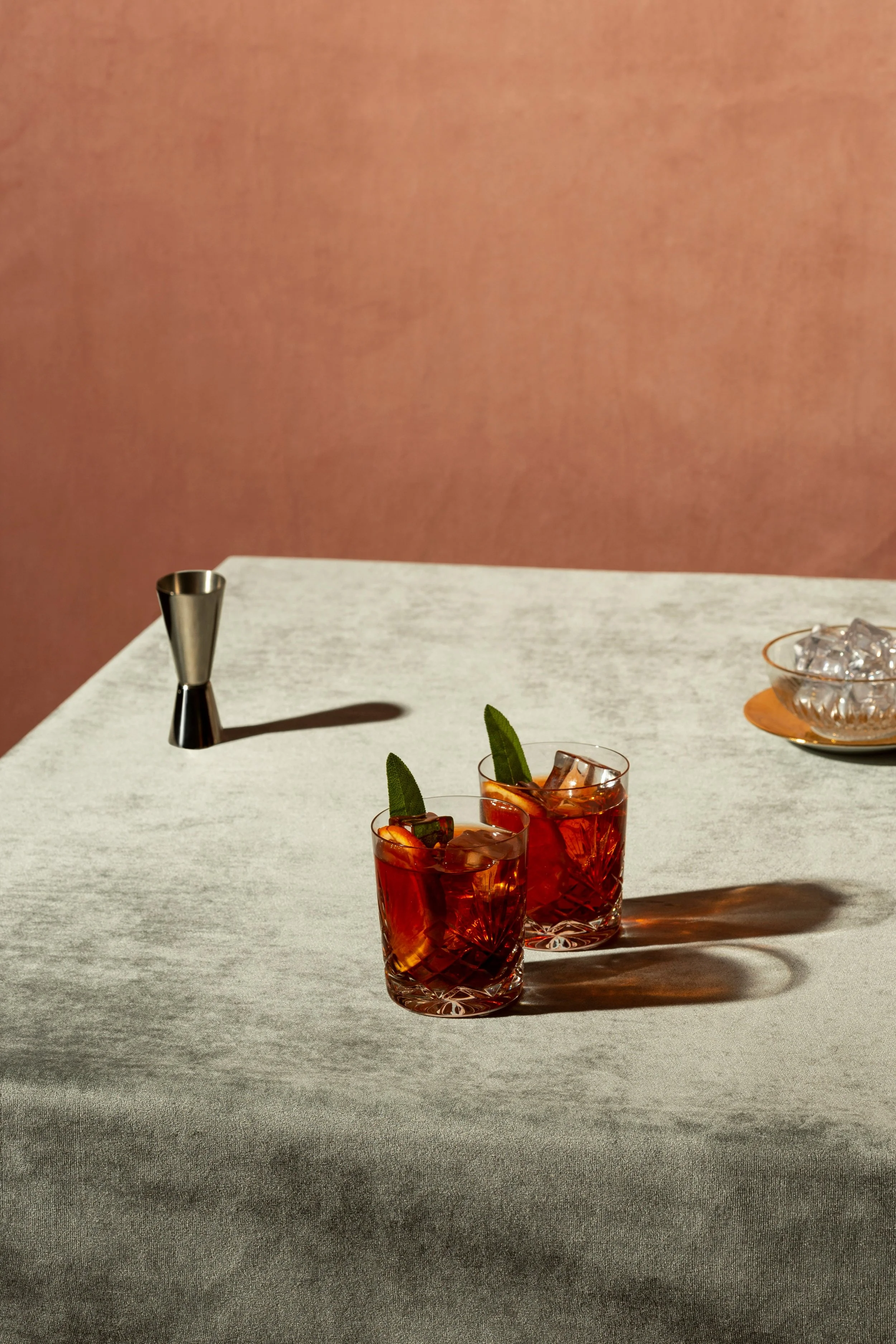 To røde drinker med is og sitronbiter på en hvit bordduk, en metallkopp og en glasskål med is i på en rosa bakgrunn.