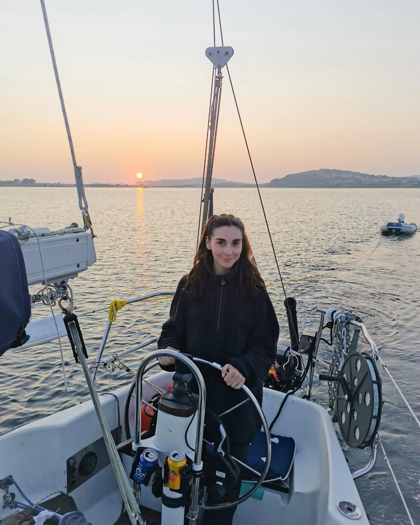 En kvinne som står ved roret på en seilbåt i solnedgangen, med vann og fjell i bakgrunnen.