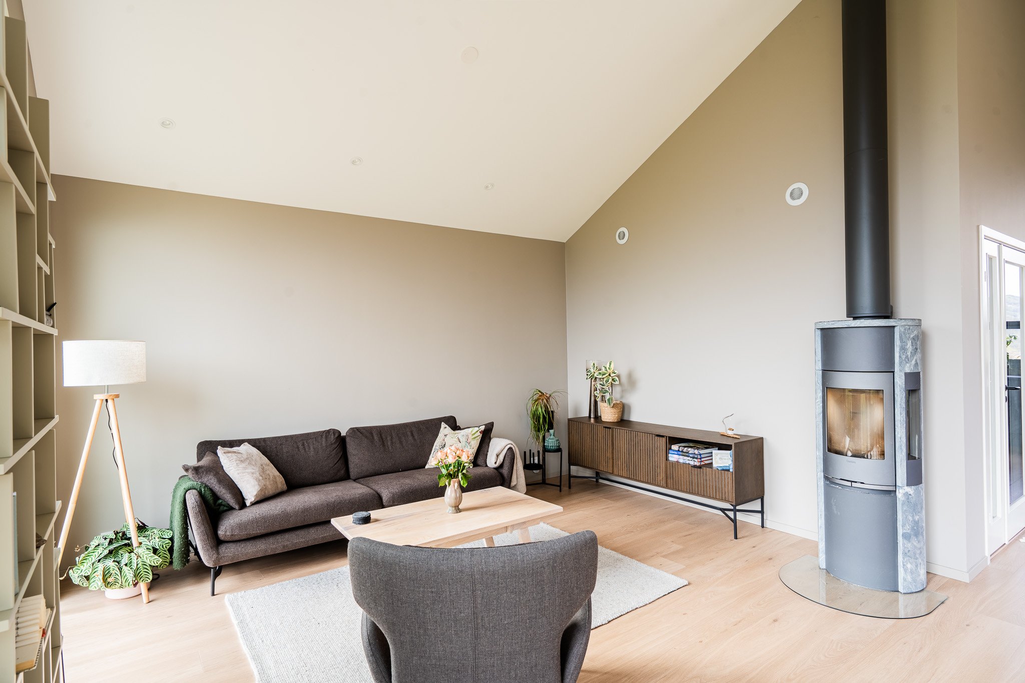 Stue med sofa, spisebord, peis og bokhyller, med store vinduer og planter.