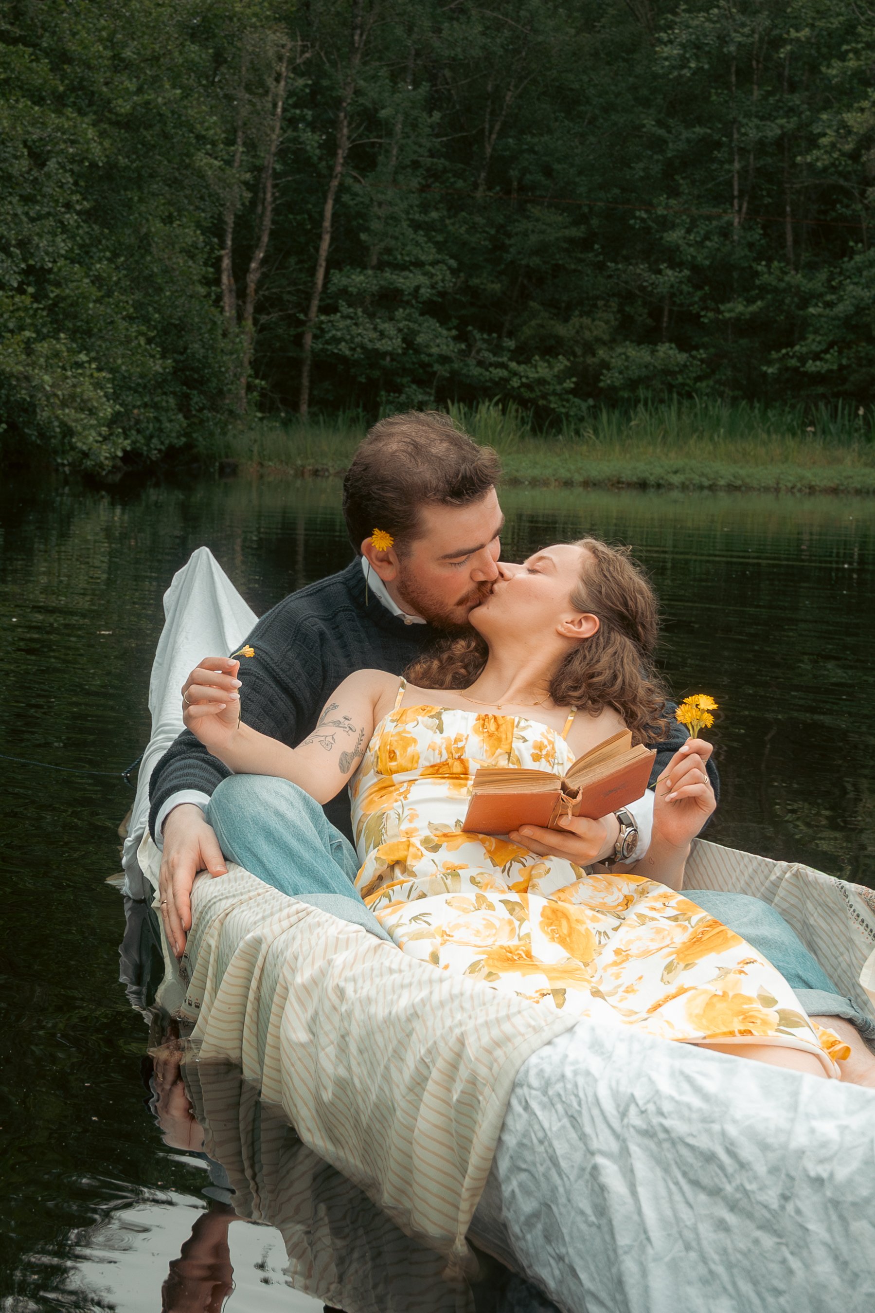 Et par kyssende personer i en båt på en innsjø, med en åpen bok og blomster i hendene.