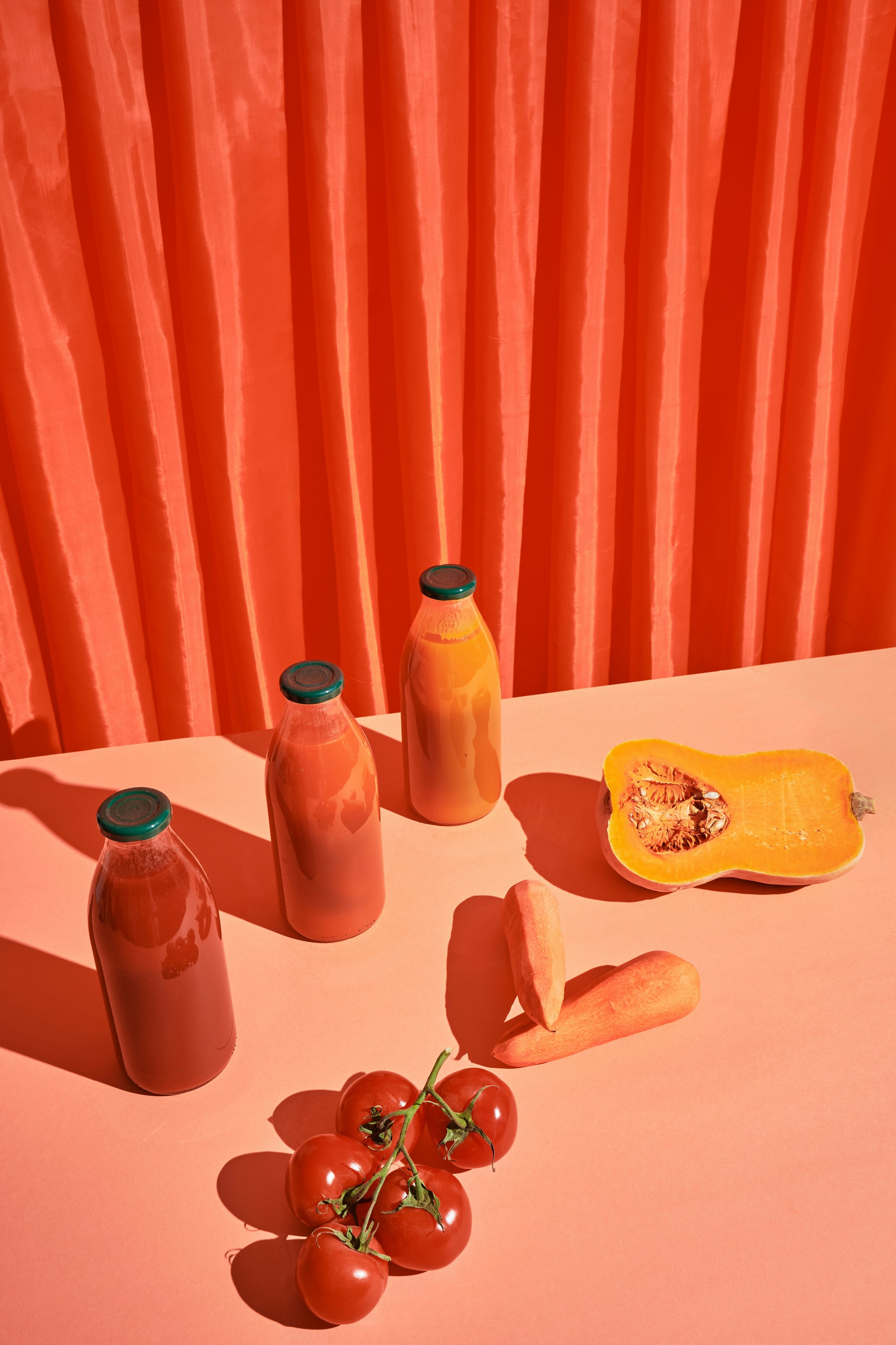 Bilde av tre flasker med oransje drikke på en bordplate, en halv oransje gresskar med frø og kjøtt, en klase cherrytomater, en gulrot, to gulrøtter, og en sitronbåt, mot oransje gardiner.