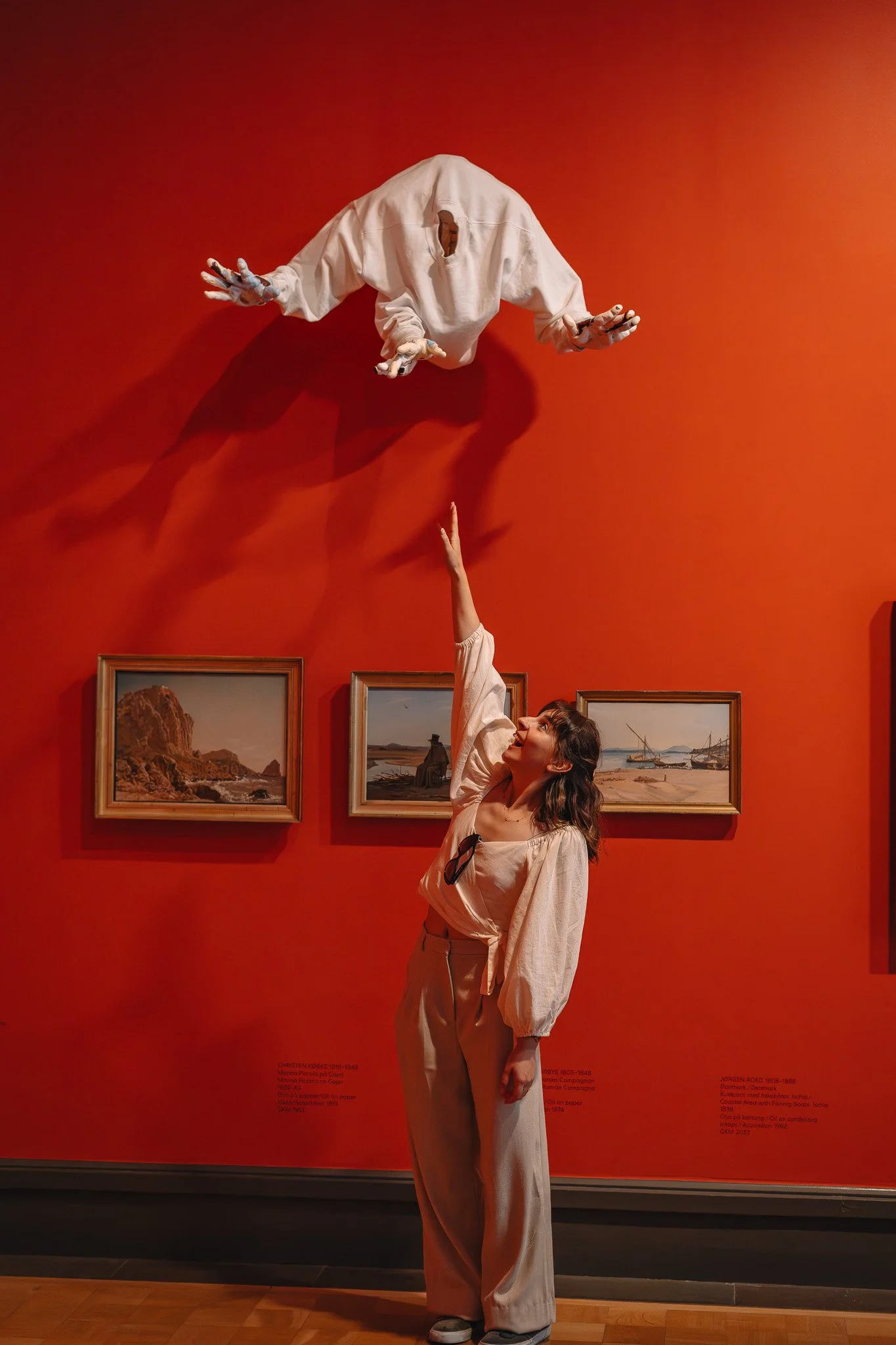 En kvinne står i et rom med rød vegg og tre malerier, og tar tak i en opphengt uttrykkende mannsskulptur. Skulpturen har utstrakte armer og et hull der hodet skal være.