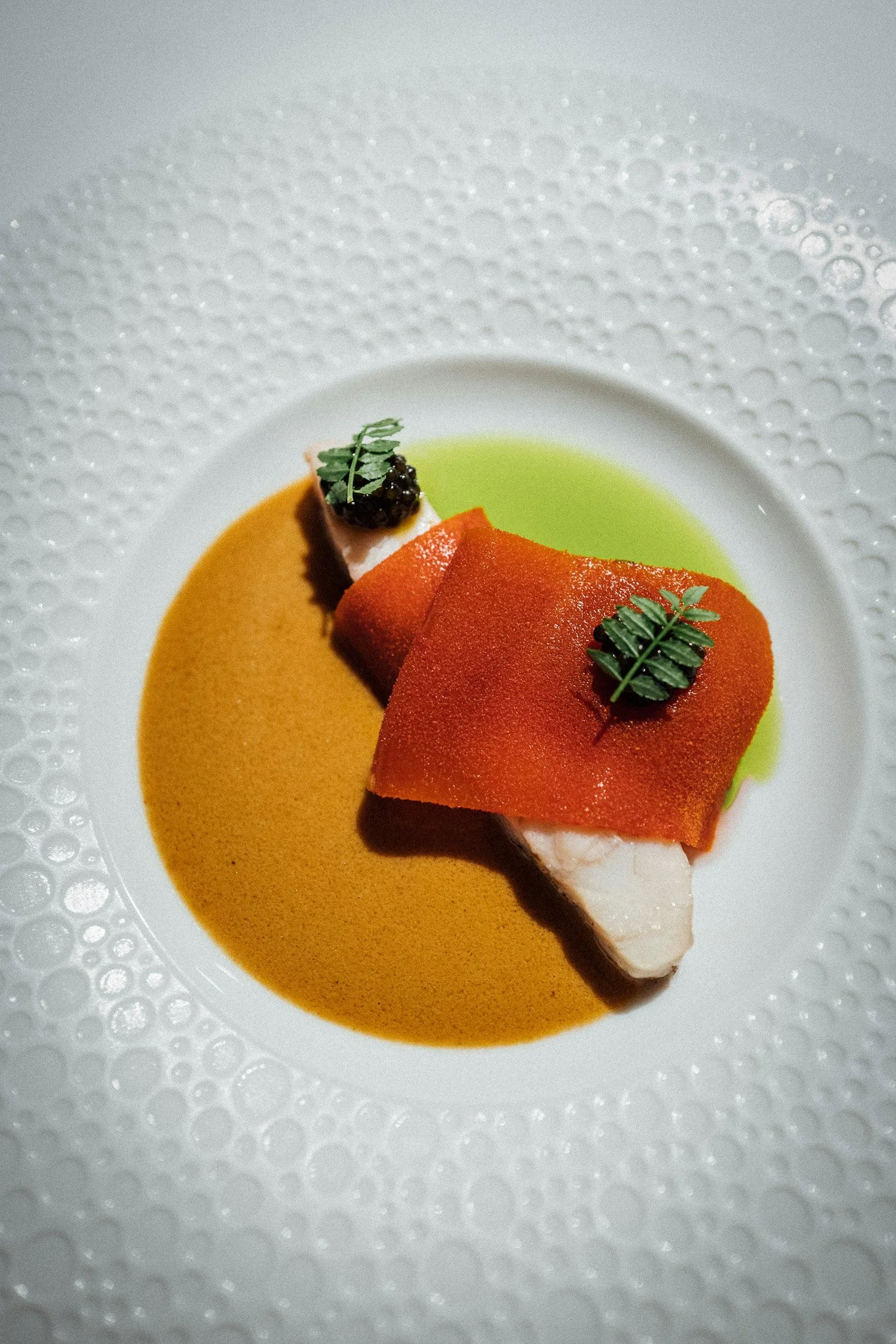 En tallerken med en rett, dekorert med grønne blader, omgitt av en oransje saus og en grønn saus, servert på et hvitt fat med en mønstret kant.