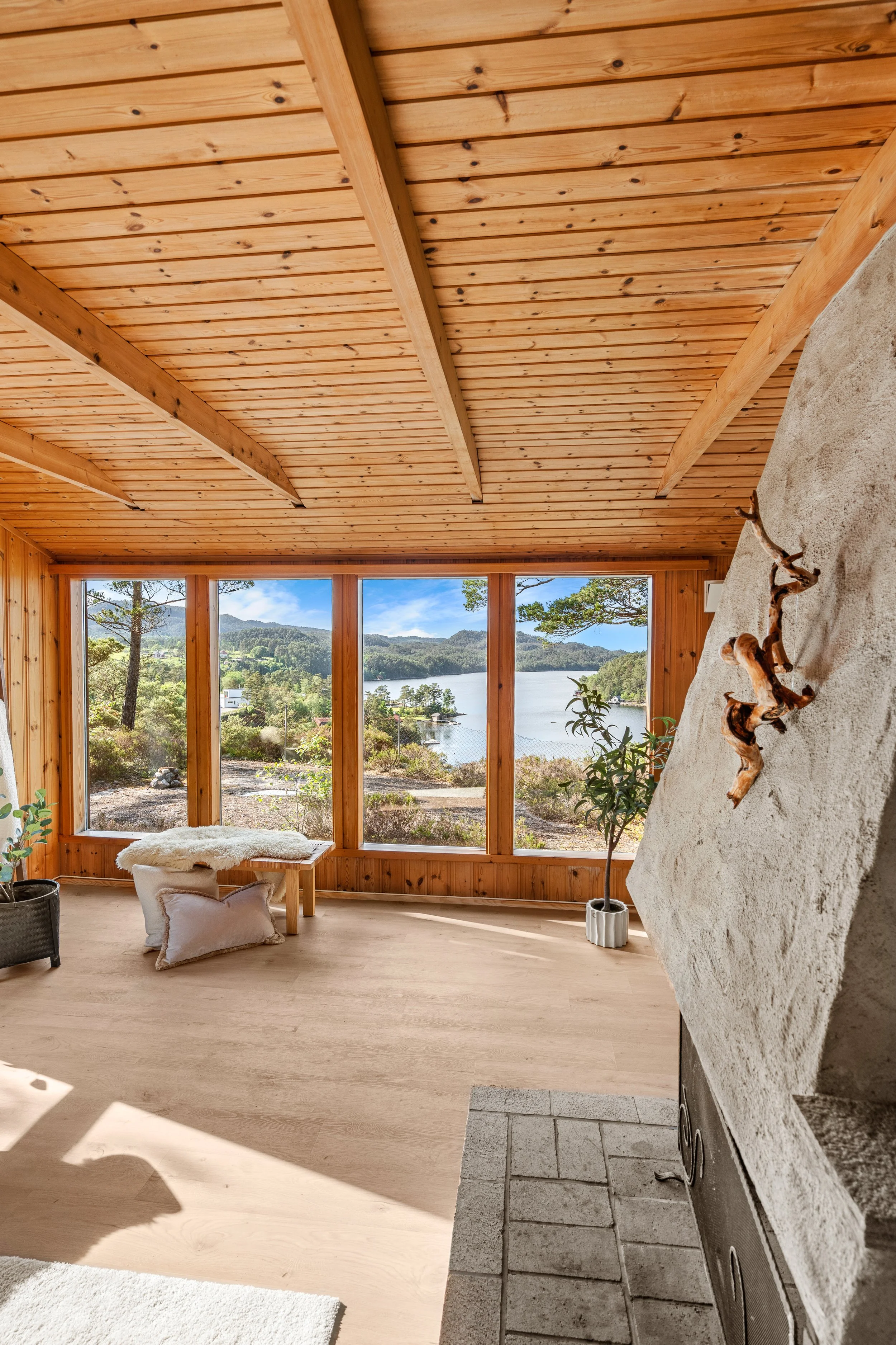 Innendørs område foran store vinduer med utsikt over innsjø og fjell, trepanel i tak og vegger, sofa med pelsdekket ottoman, pynt med grønne planter og dekorativ steinvegg med peis.