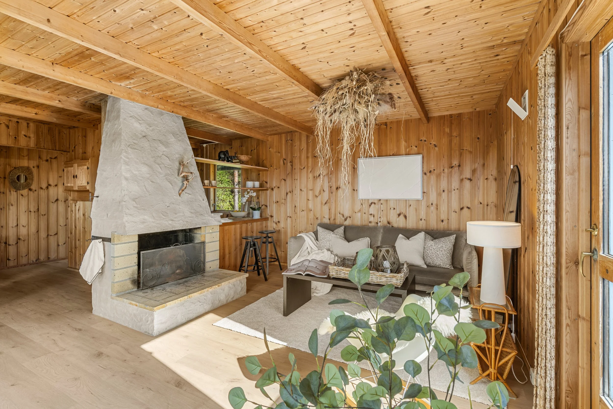 Innendørs bilde av et rom med trepanel på vegger og tak, en peis, en sofa med puter, et lite bord med en åpen bok, og grønne planter, med store vinduer og naturlig lys.