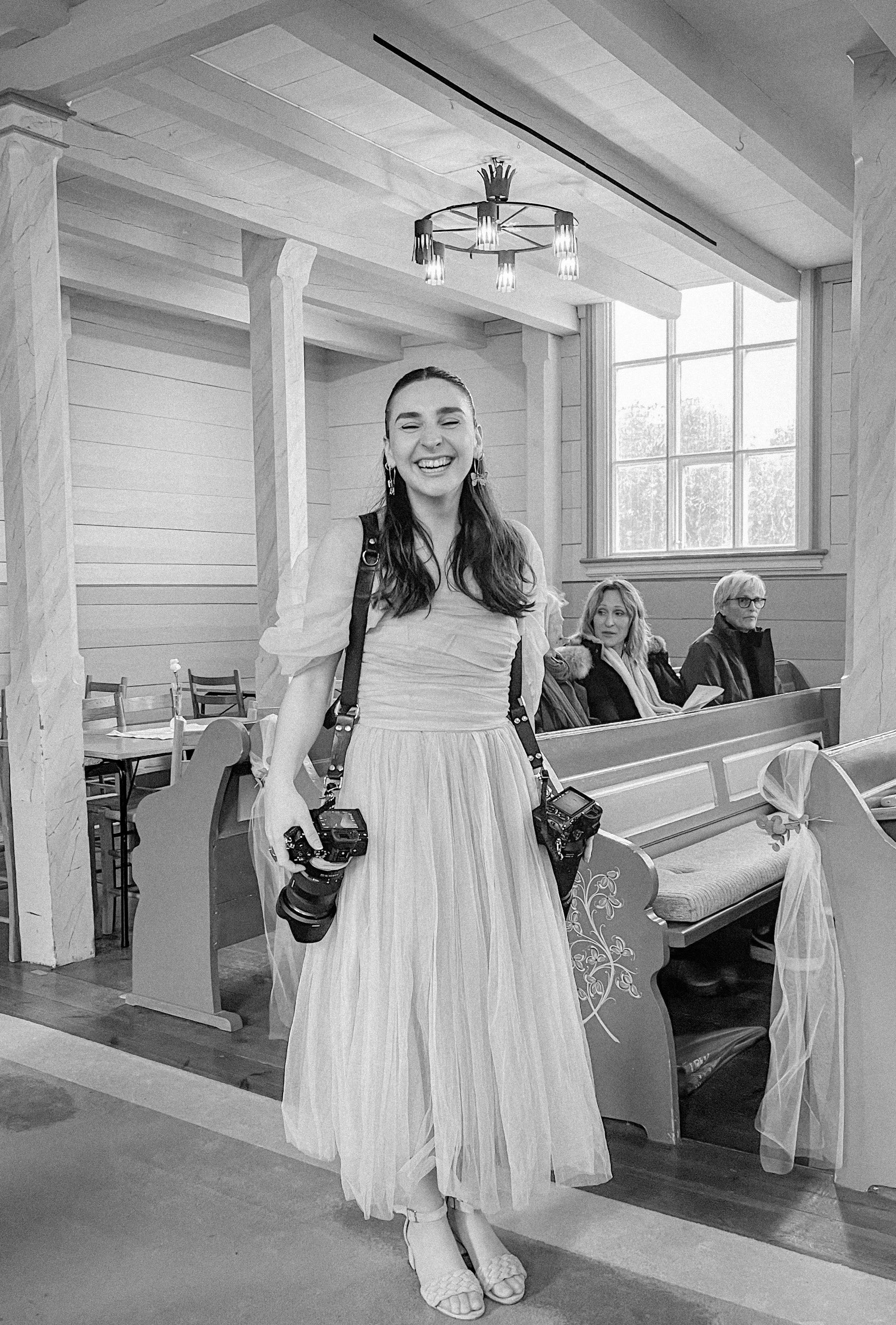 En kvinne i lang kjole som smiler, med to kameraer hengende rundt halsen, inne i en kirke eller en gammel bygning. To kvinner sitter foran i benkeseksjonen i bakgrunnen.