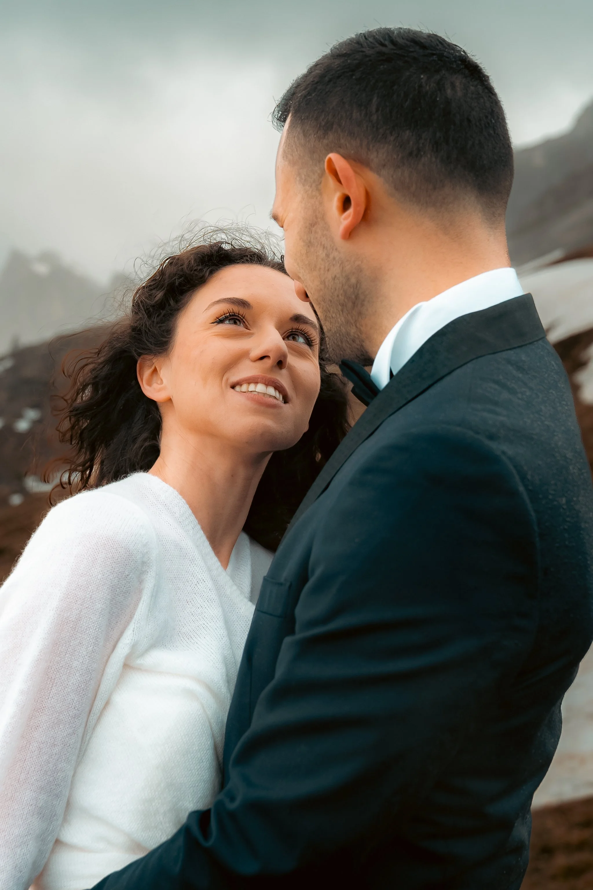 Et romantisk øyeblikk mellom en mann i smoking og en kvinne i en hvit genser, utendørs med fjell i bakgrunnen.