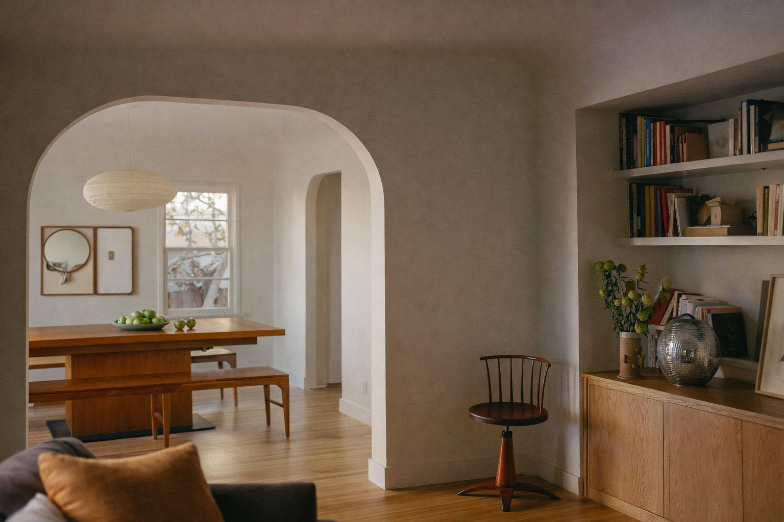 Innendørs rolig boligrom med en bokhylle, vaser og malerier, sett gjennom en arkadearm. Det er naturlig lys, varmt treverk og et avslappet atmosfære.