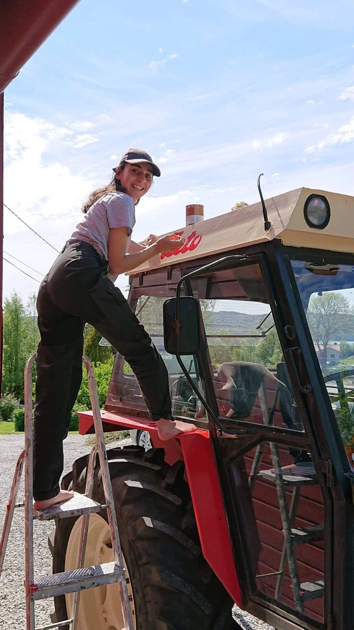 En kvinne står på en trappestige og maler en tractor utendørs under en klar dag.