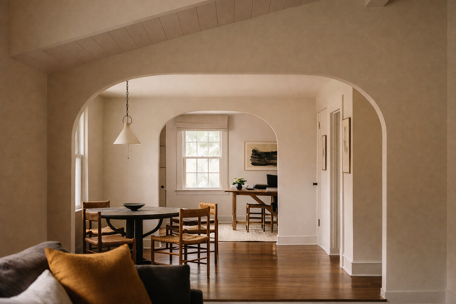 Innendørs bilde av et rom med spisebord, stoler og skrivebord, med store vinduer som slipper inn naturlig lys.