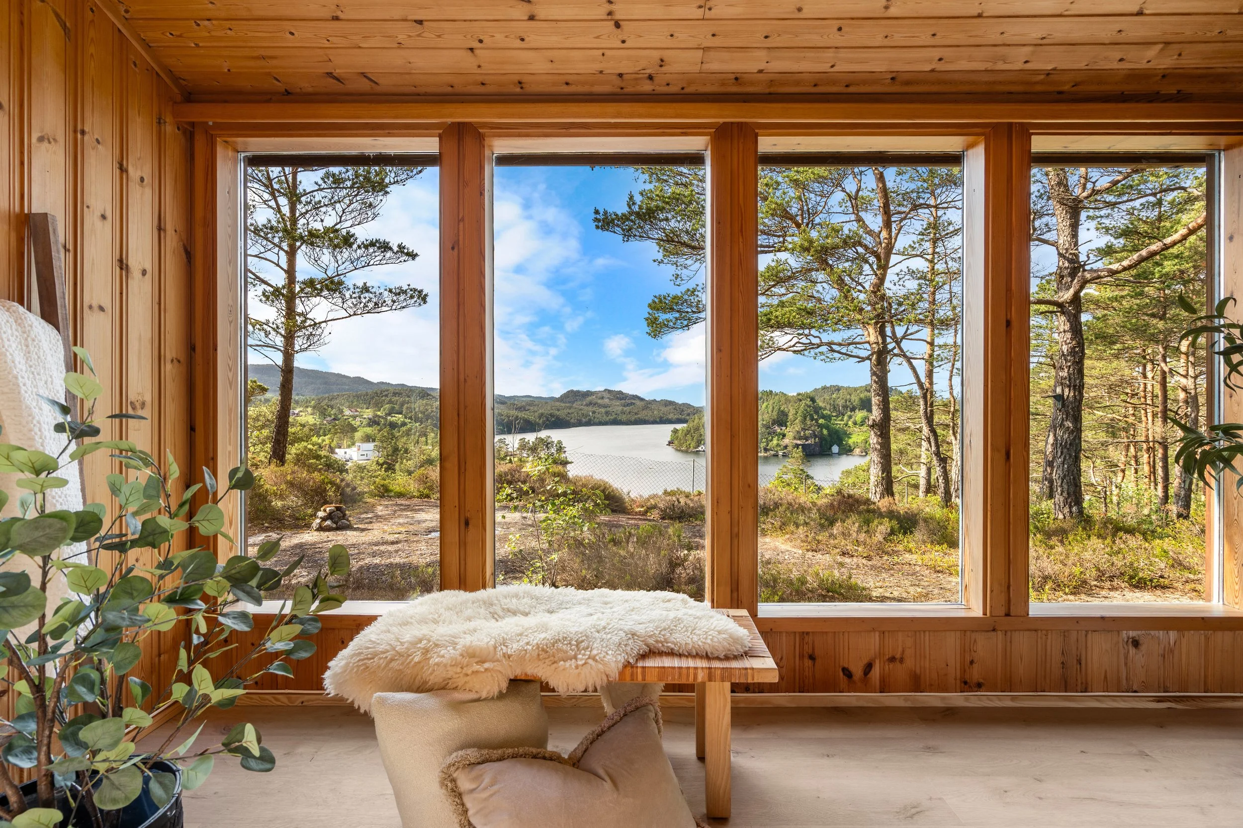 Innendørs rom med store vinduer som viser utsikt over skog og fjell, trestenvægger, et fluffy vek, og en puf, del av et koselig rom.