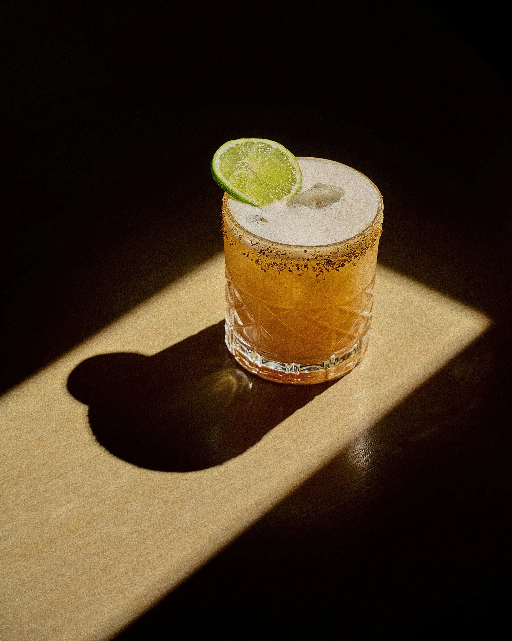 Et this glass med en limebåt og isbiter, med salt rundt kanten, står på et lyst bord. Rødt lys reflekteres fra glasset.