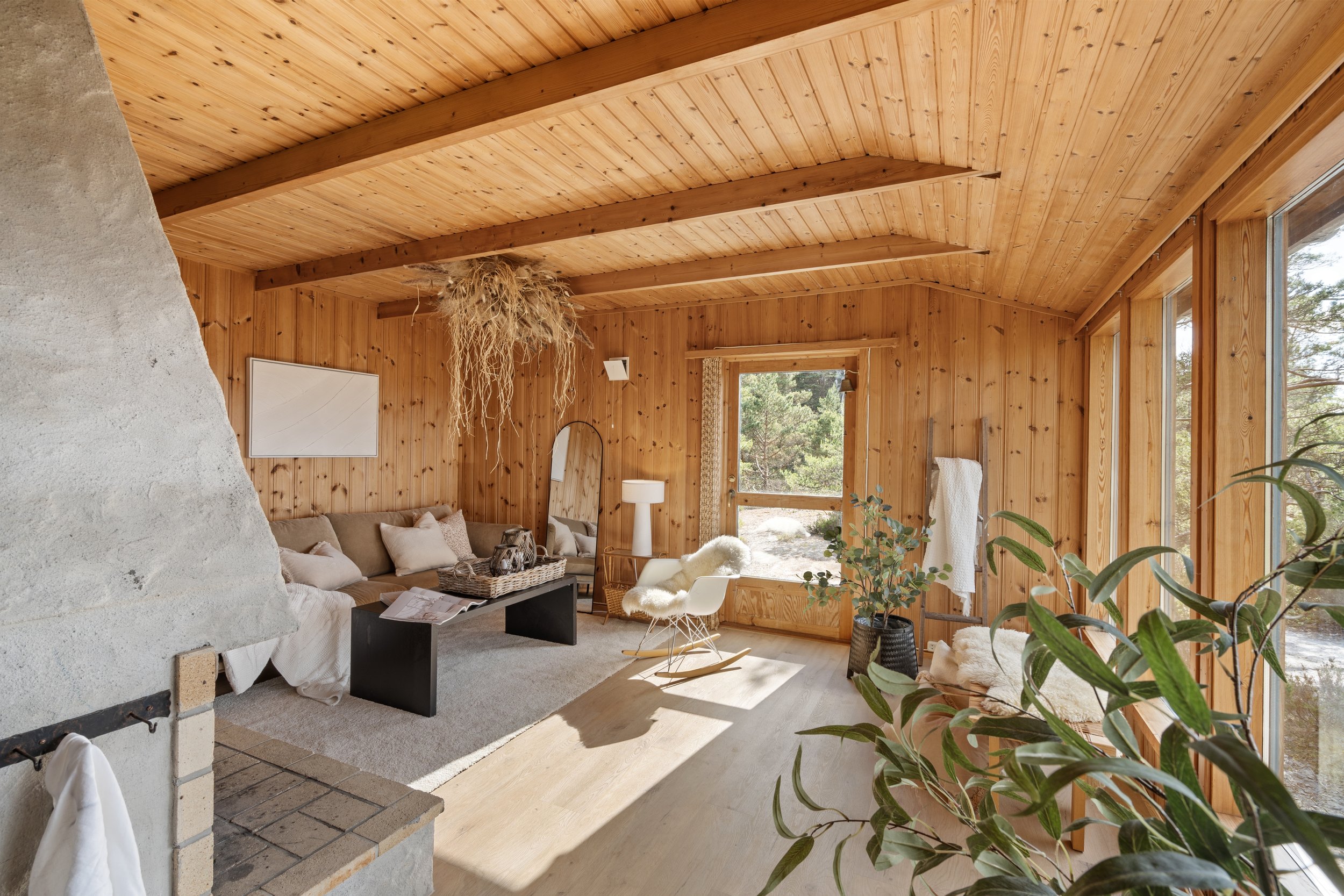 Innendørs stue med trevegger, store vinduer og en takdekk av tre. Møbler inkluderer sofa, salongbord, lamper og lenestol, med planter og dekorasjoner, samt en peis.
