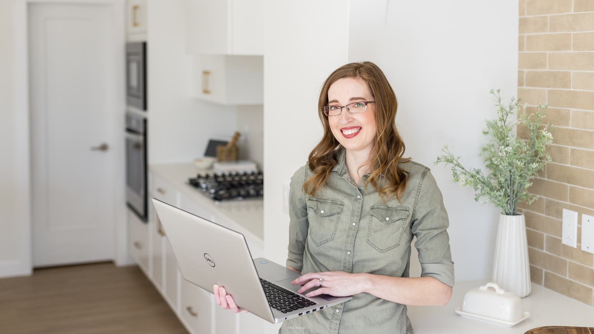 A woman with glasses and an olive green shirt smiling and holding a laptop in a bright, modern white and wood kitchen.
