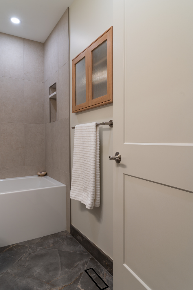 A side view of a contemporary bathroom with dark hexagonal floor tiles and large format wall tiles. A glass shower door is visible on the right, next to a wood-framed wall cabinet, a hanging towel, and a white bathtub.