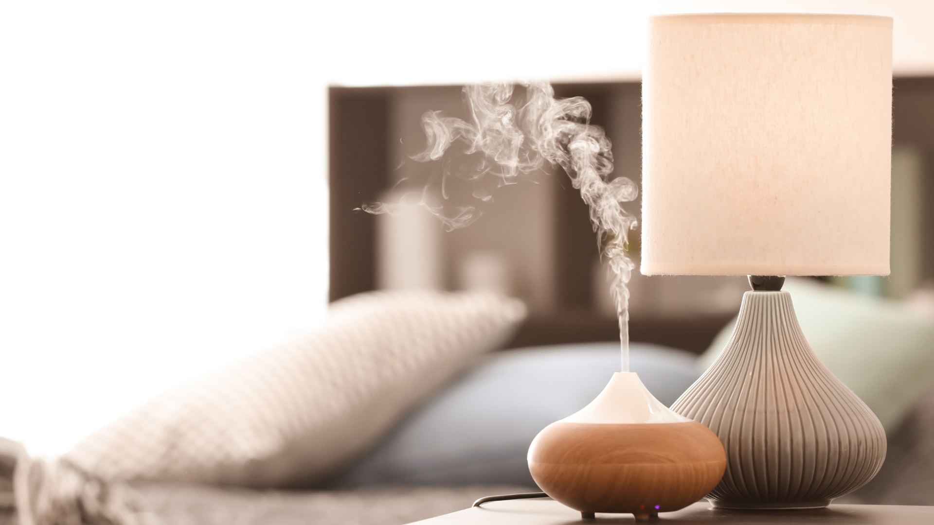 Modern bedroom with an essential oil diffuser emitting calming steam beside a textured ceramic table lamp on a nightstand. Soft, neutral bedding in the background creates a relaxing, spa-inspired atmosphere.